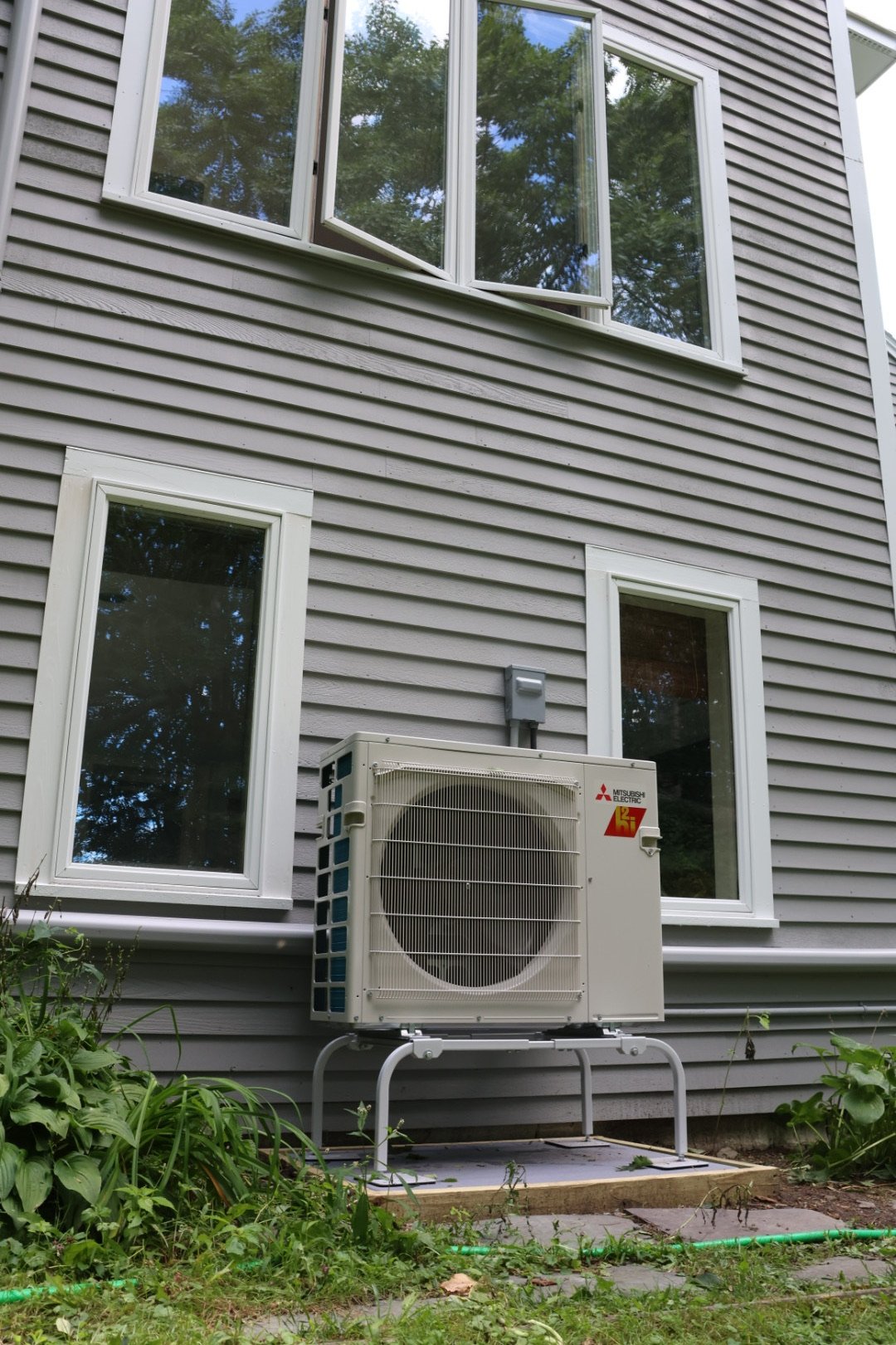 Mitsubishi Electric outdoor heat pump on a raised stand beside a gray-sided home.