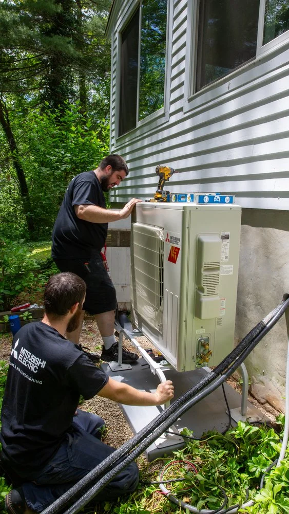 Two HVAC technicians from Only Heat Pumps installing a Mitsubishi Electric outdoor heat pump unit beside a home in Southern Maine.