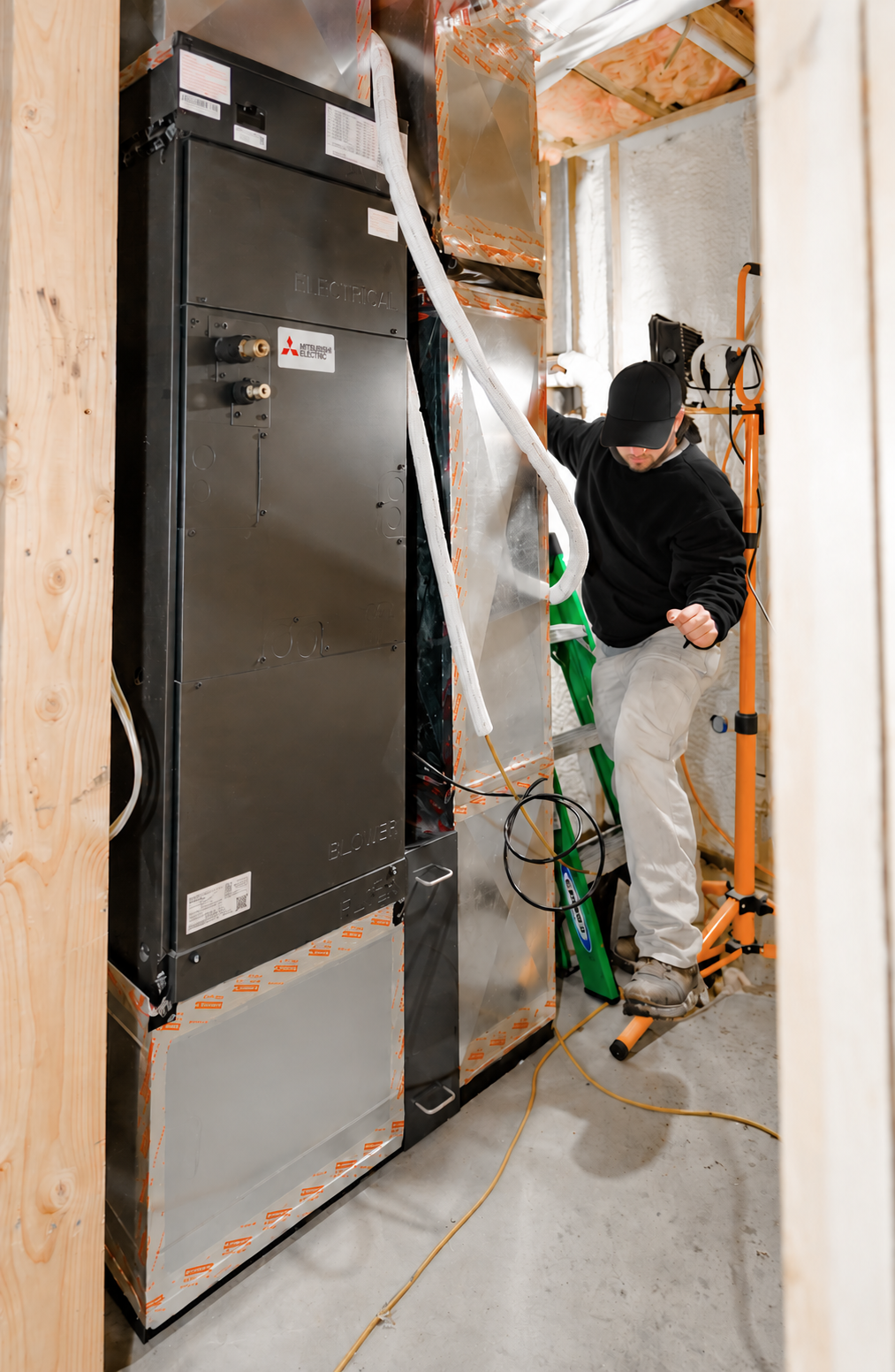 HVAC technician wearing a black uniform working beside a Mitsubishi Electric ducted heat pump system in an unfinished mechanical room.