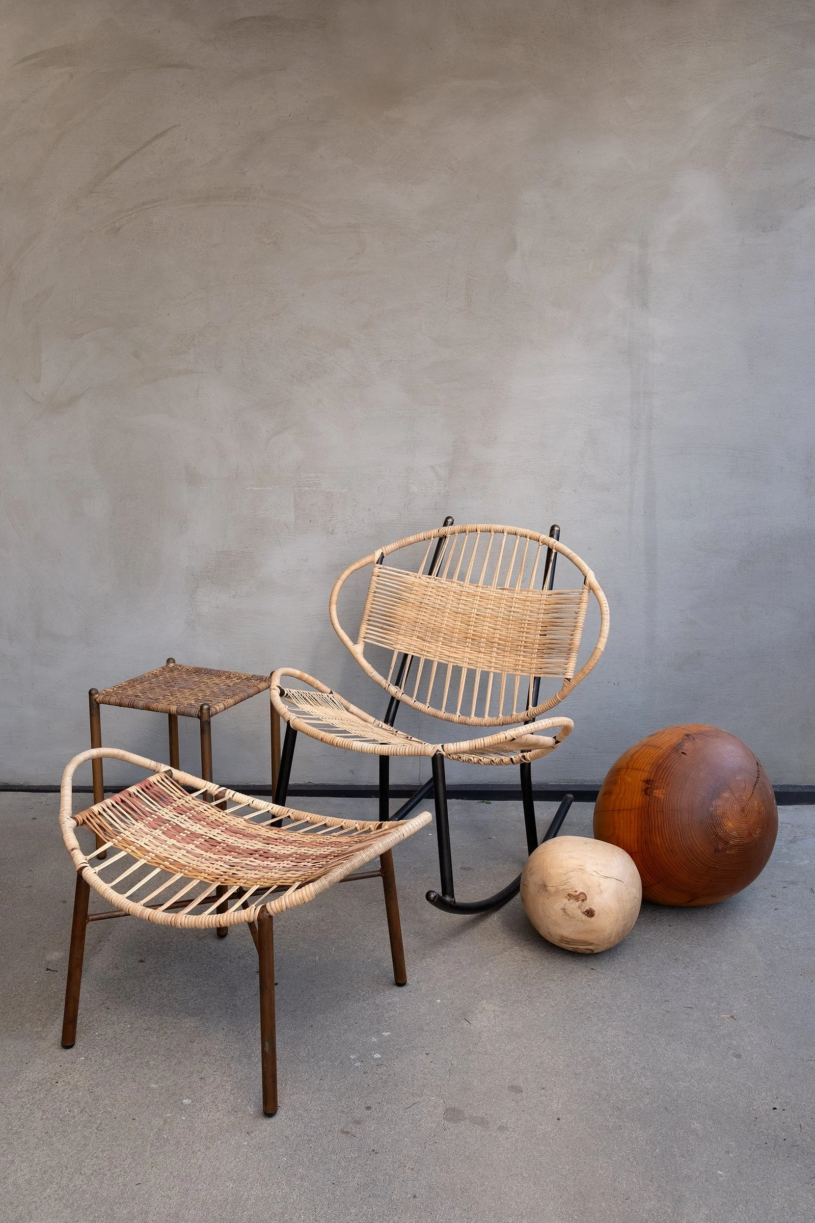 Clamshell rocking chair with woven cane seat, black steel frame, and matching ottoman. Styled with small wooden spheres on a concrete floor against a textured wall.