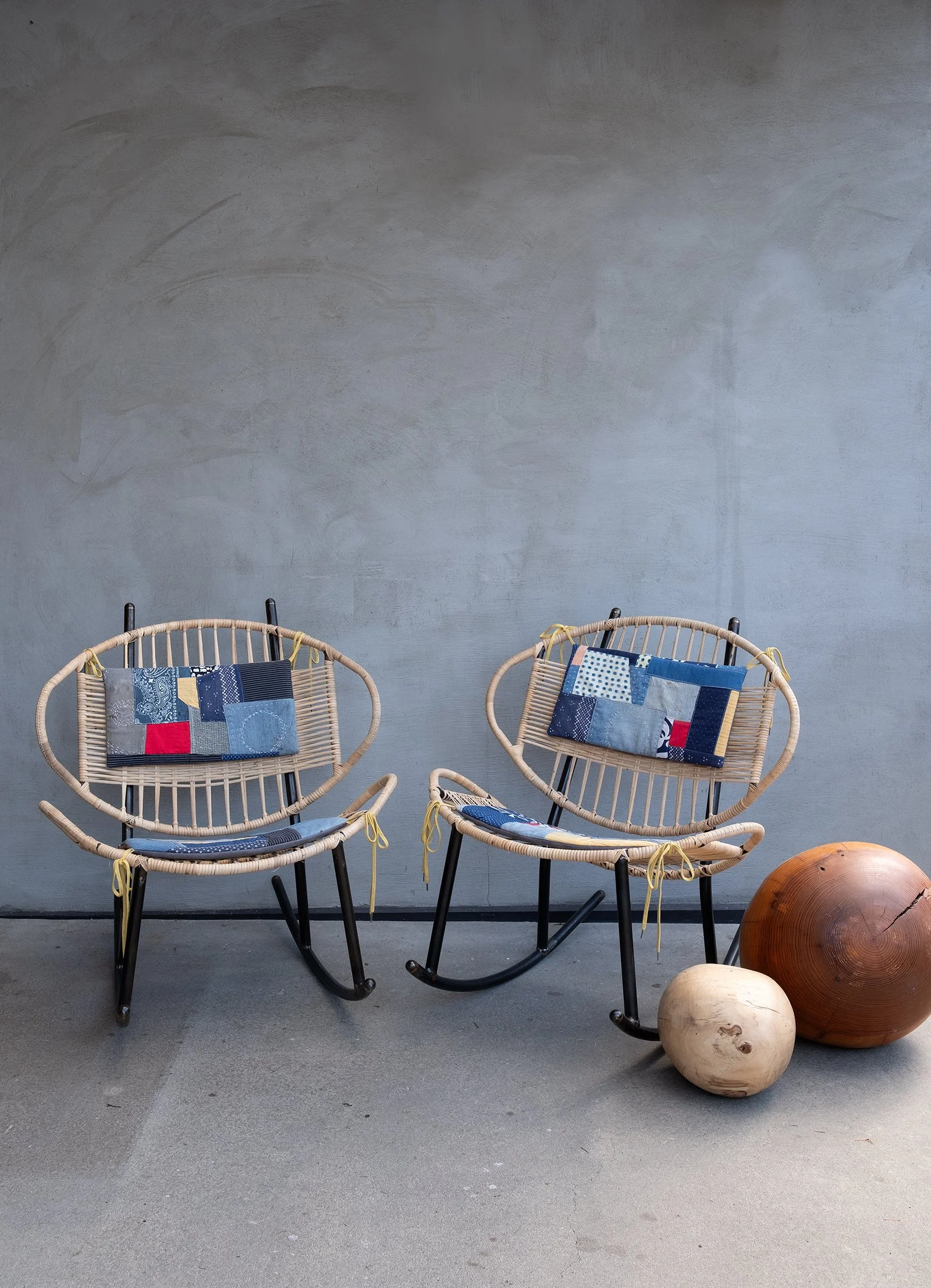 Two rattan Clamshell rocking chairs with colorful Sashiko-style woven cushions, positioned next to large wooden spheres on a concrete floor against a gray wall.