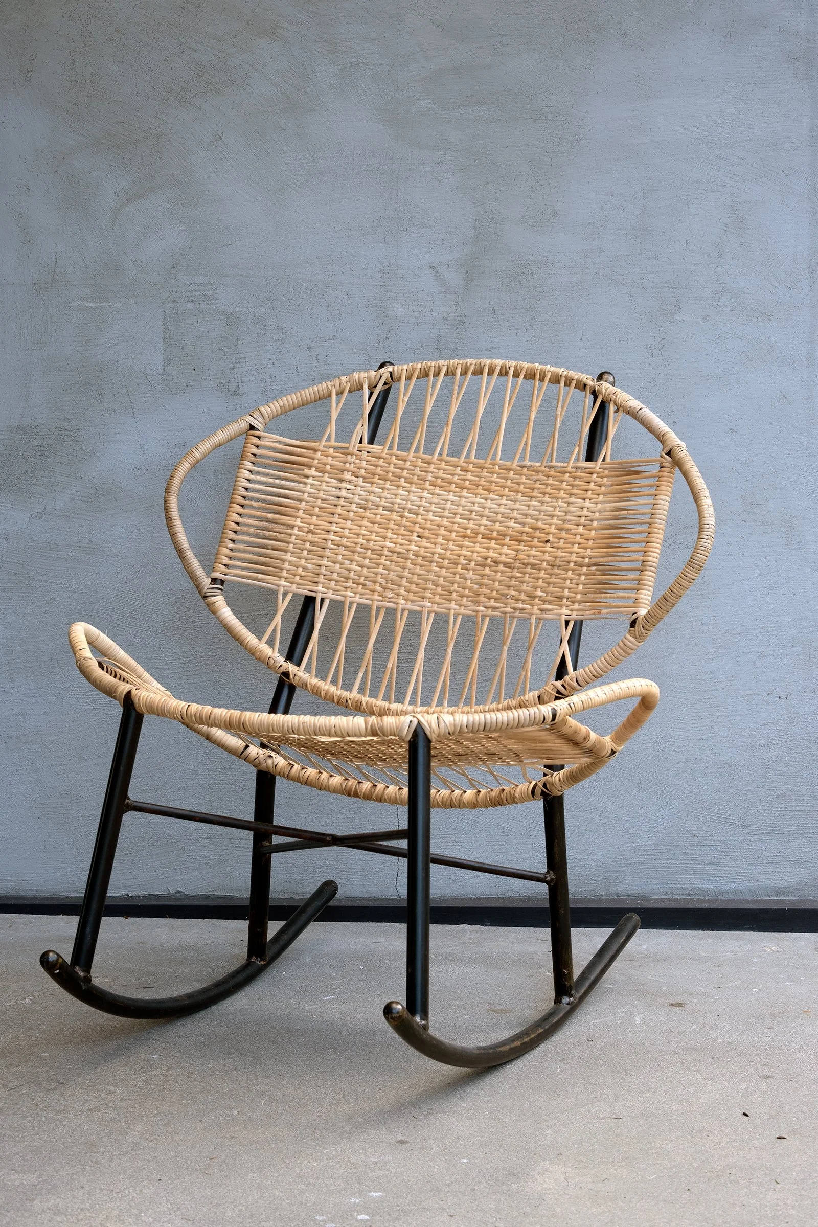 Woven cane Clamshell rocking chair with black steel frame, set against a modern gray wall for a minimalist, stylish look.