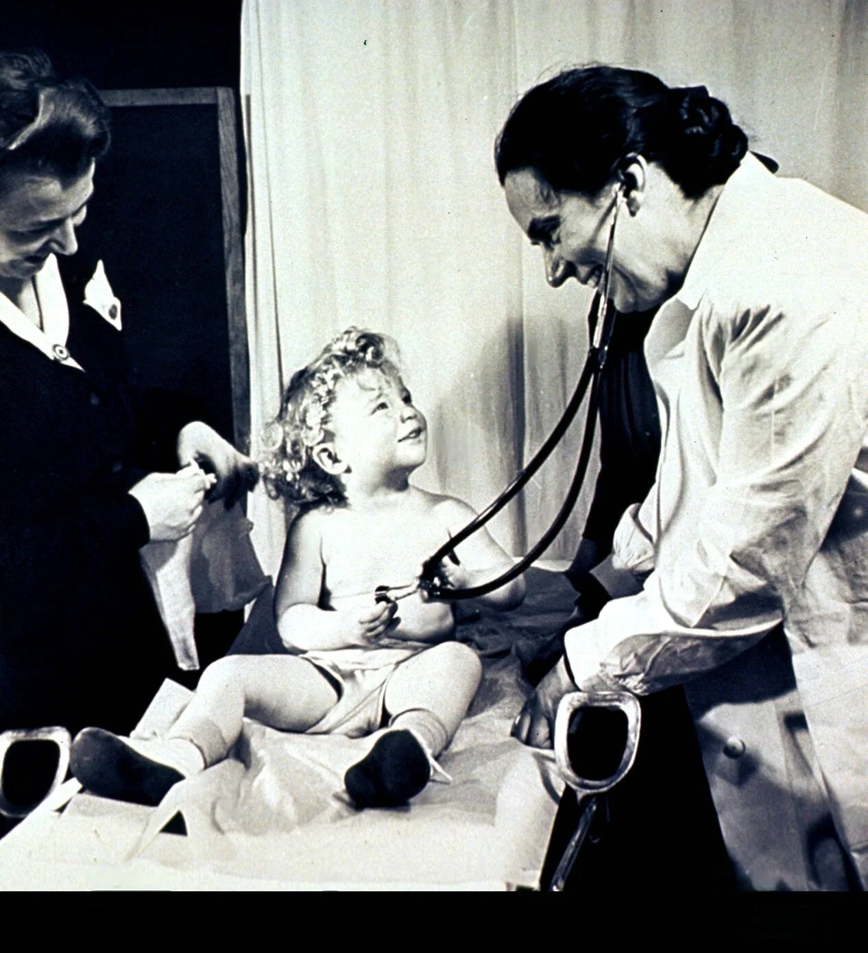 Vintage photo of a doctor examining a smiling child with a stethoscope, while another person assists in a medical setting.