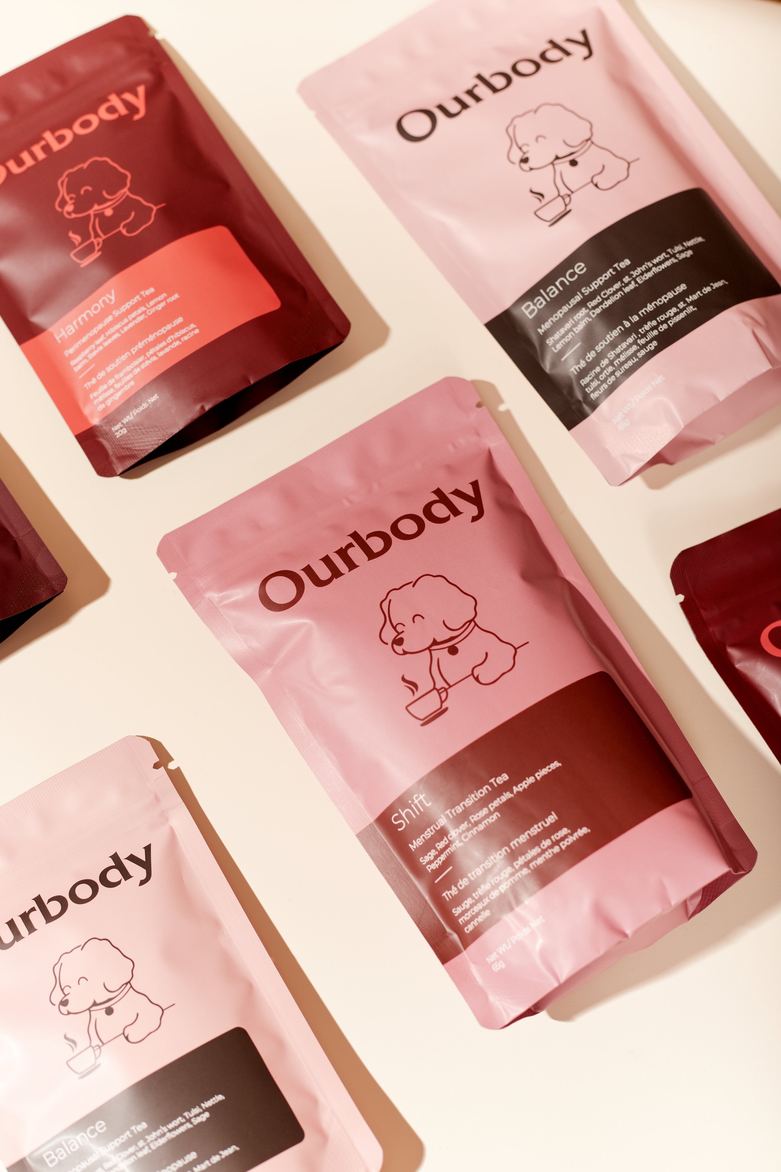Various pink and red packages of Ourbody herbal tea products featuring a cartoon dog holding a steaming mug, with names like Harmony, Balance, and Shift.