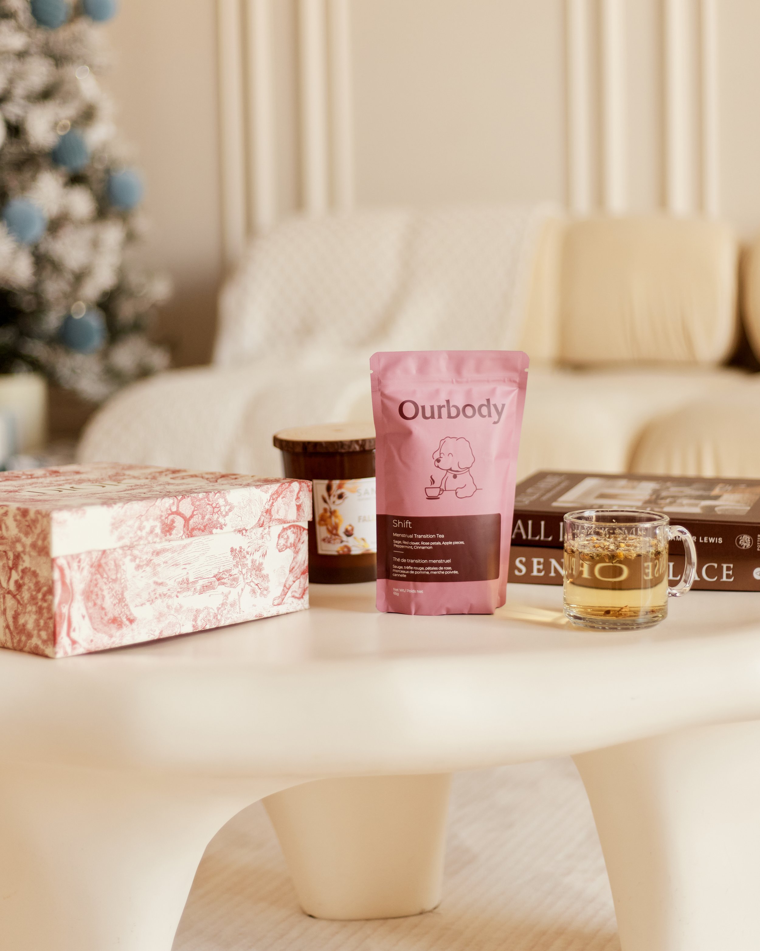 A white table with a pink package of Ourbody Shift menstrual transition tea, a cup of tea, a stack of books, a pink patterned box, and a candle, with a cream sofa and a decorated Christmas tree in the background.