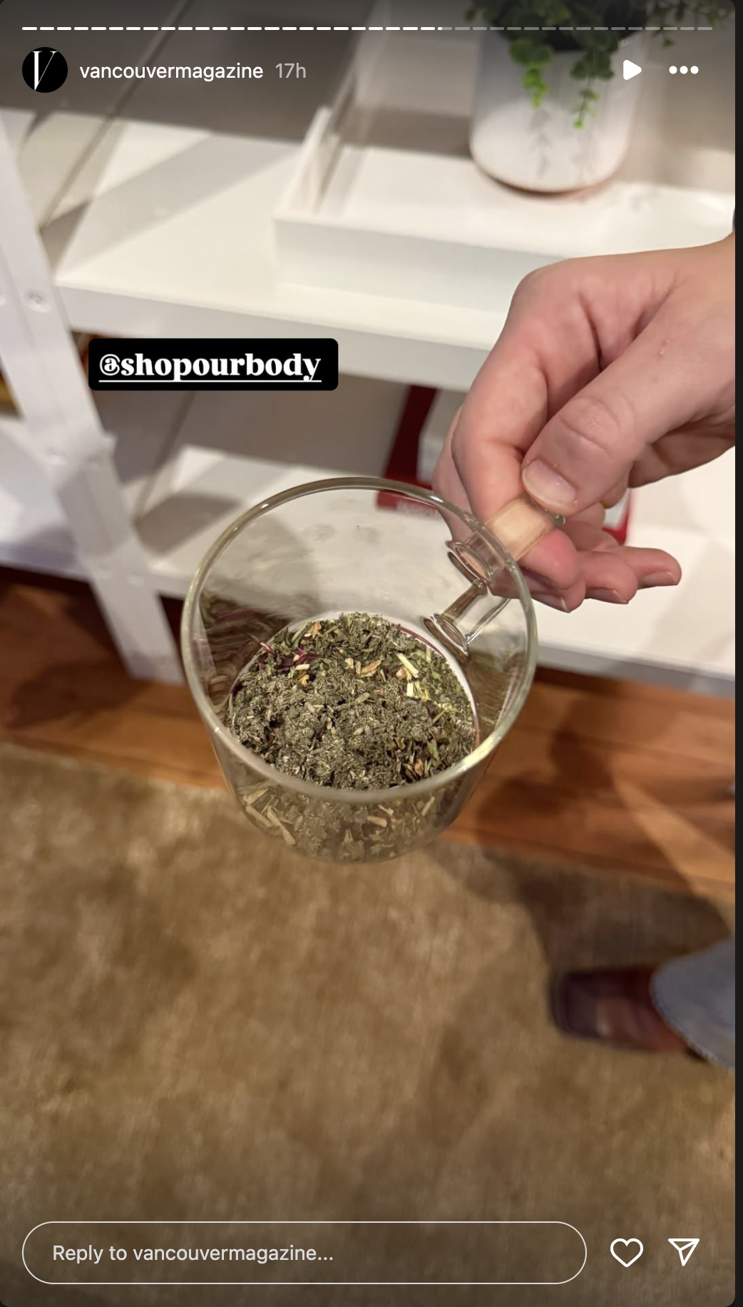 Hand holding a glass cup with dried herbal tea or loose leaf tea, with a white shelf and potted plant in the background.
