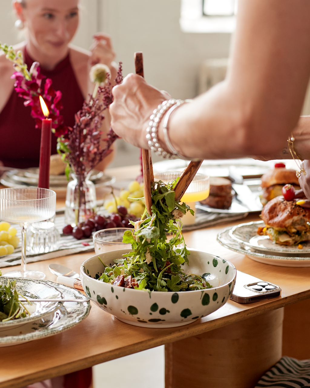 Person serving salad at a family dinner with candles, grapes, and decorated dishes on a wooden table.