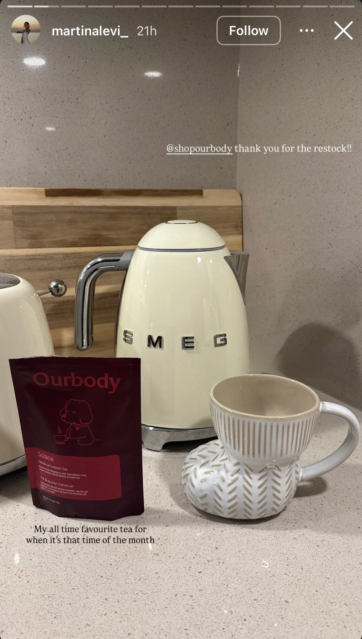 A kitchen countertop with a cream SMEG electric kettle, a striped mug with a matching saucer, and a bag of Ourbody herbal tea, with a wooden backsplash and gray walls.