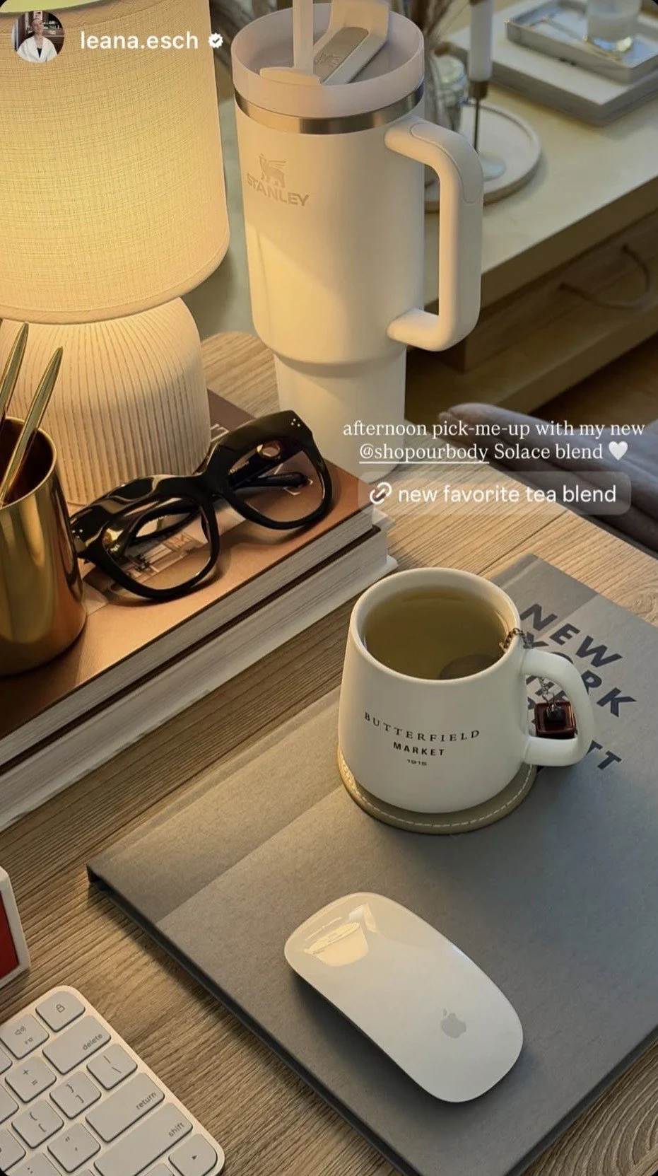 A wooden desk with a white Stanley mug filled with tea, a white Apple Magic Mouse on a gray document folder, a pair of black glasses, a closed book, a metallic gold cup with pens, a cream-colored lamp, a large white Stanley thermos, and a blurred background of a room with decorative items.
