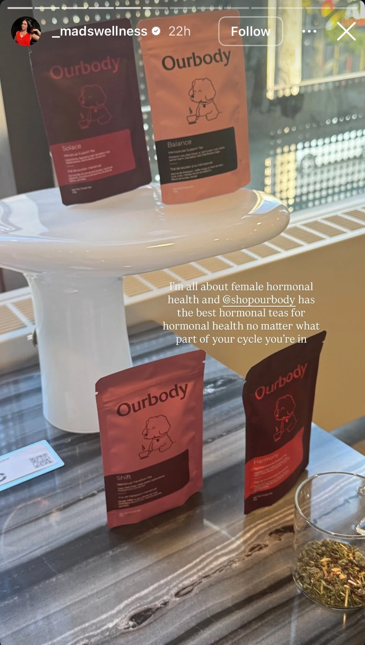 Displays four packets of Ourbody herbal teas on a white stand and table, with a bowl of herbal tea ingredients nearby. A person’s Instagram message overlay highlights the teas as beneficial for female hormonal health.