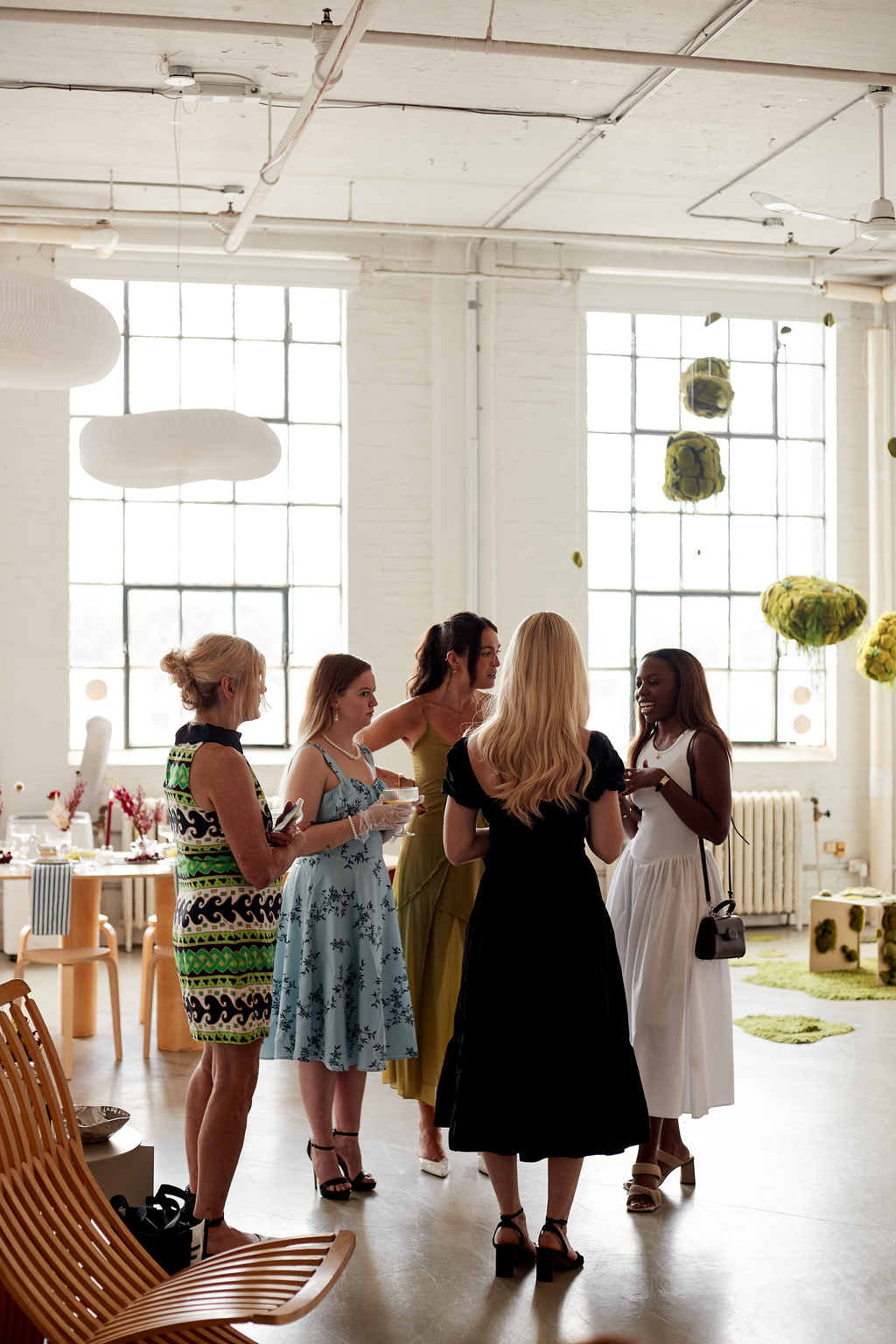 Five women in colorful dresses standing in a bright, modern indoor space with large windows, engaging in conversation.