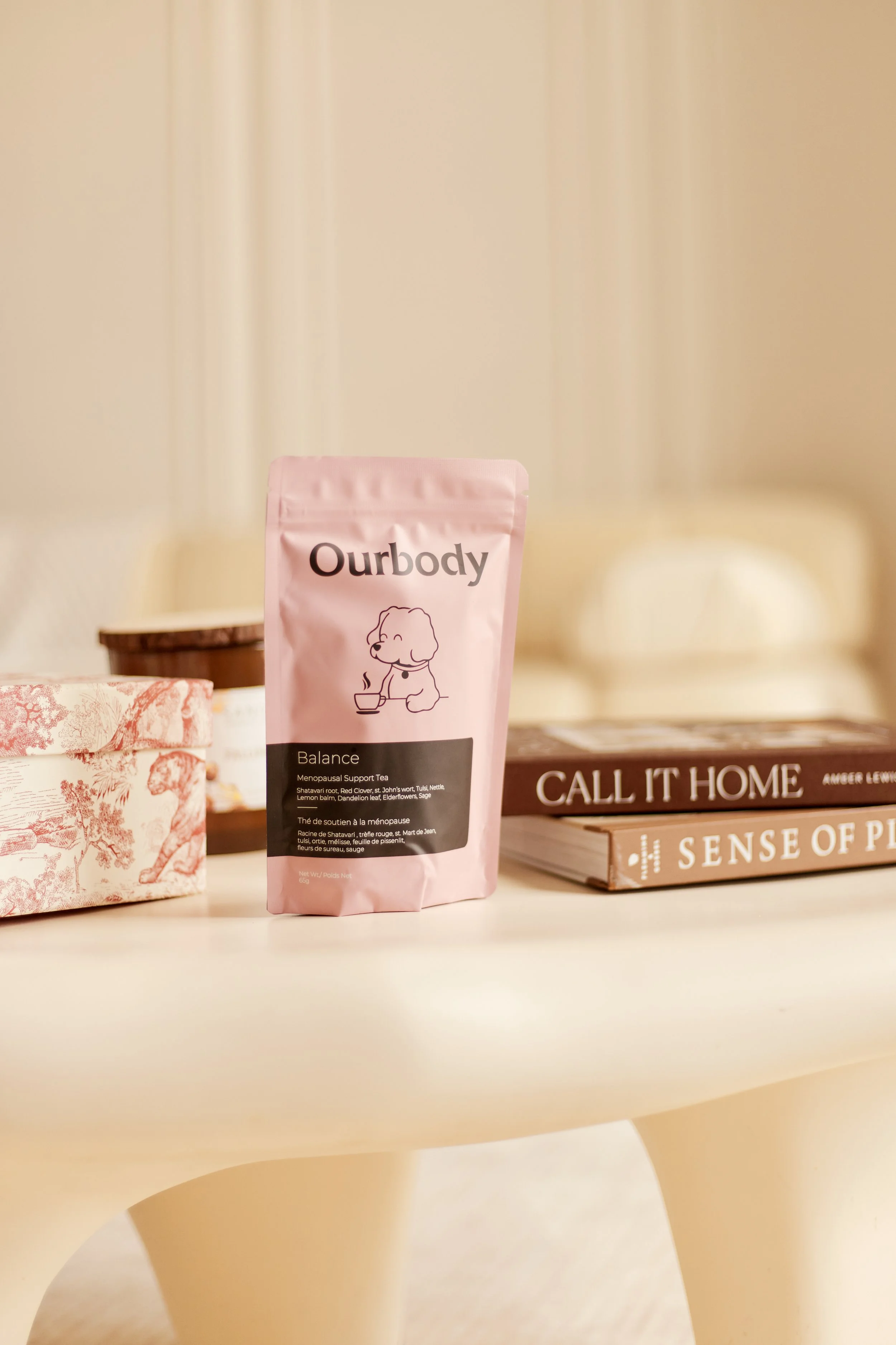 A pink package labeled "Ourbody Balance" Menopause Support Tea on a white table, with a stack of books and a decorative box nearby, in a cozy room.
