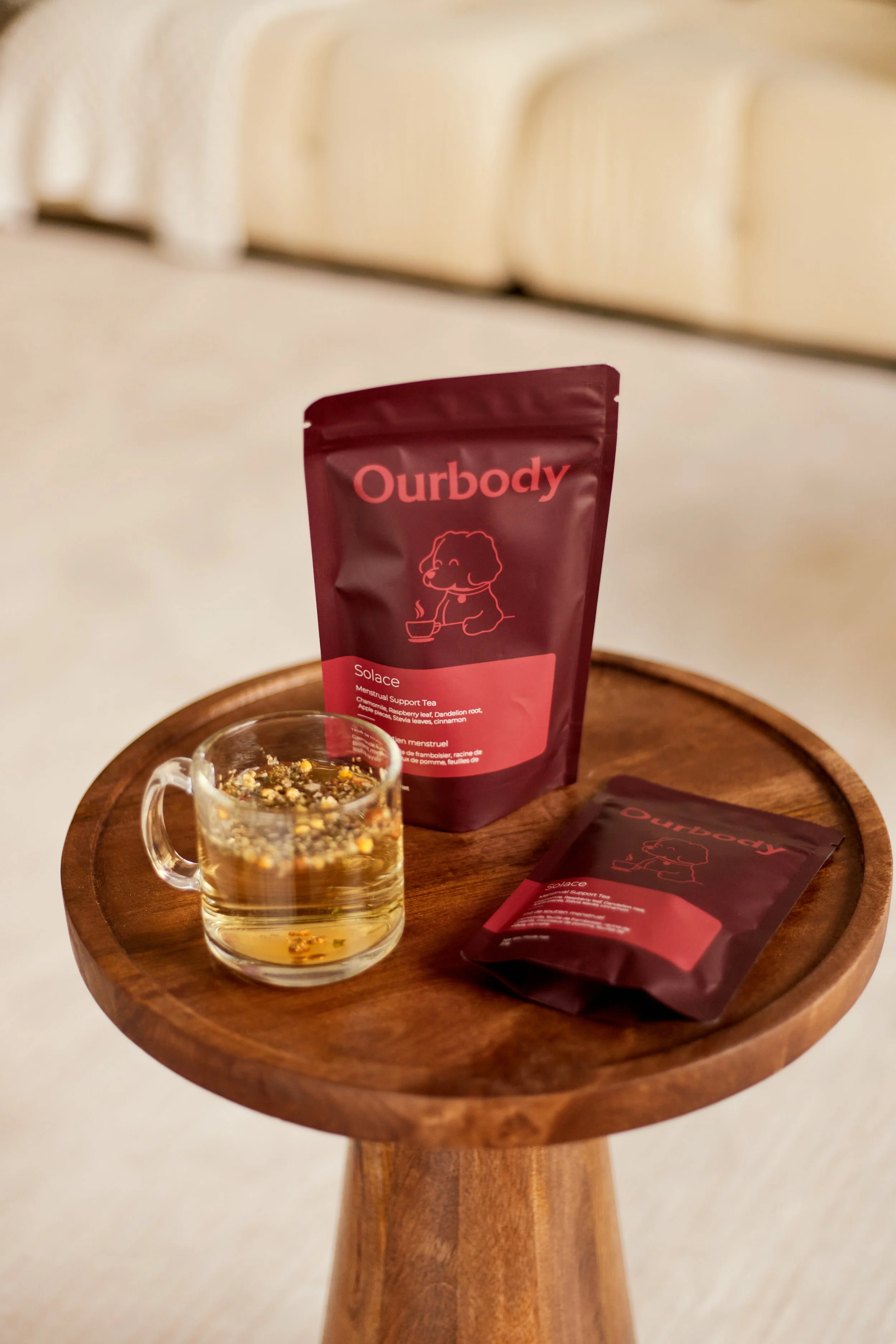 A wooden table holding a glass cup with herbal tea, a bag of Ourbody Solace herbal tea, and a smaller packet of the same tea, in a cozy indoor setting.
