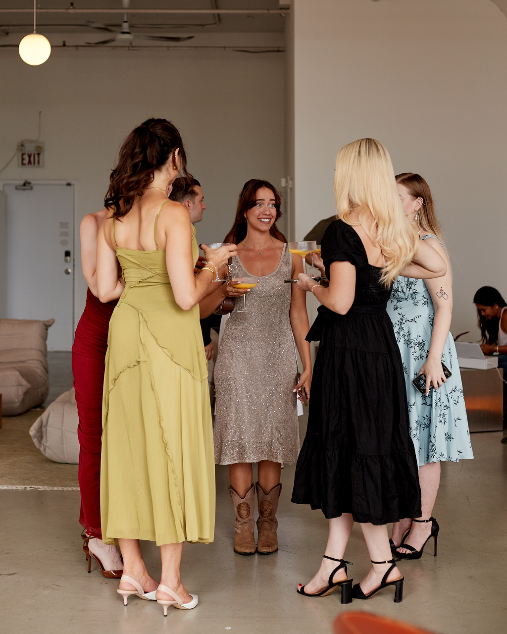 Group of women at a social event, standing and talking with drinks in hand, dressed in dresses and heels in an indoor setting.