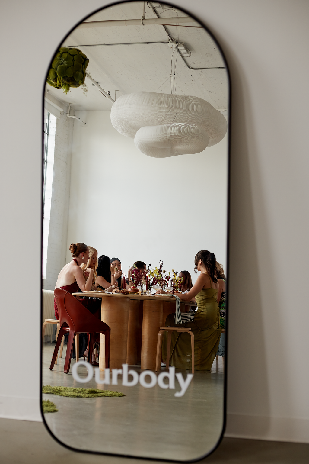 Reflection in a mirror showing a group of women sitting around a dining table with flowers in vases, in a bright, modern room with a large, white, abstract ceiling light.