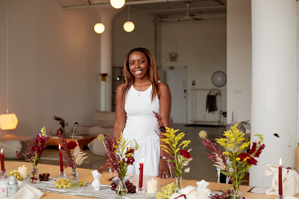 A woman standing behind a decorated dining table with candles, flowers, and wine glasses in a well-lit, minimalist room.