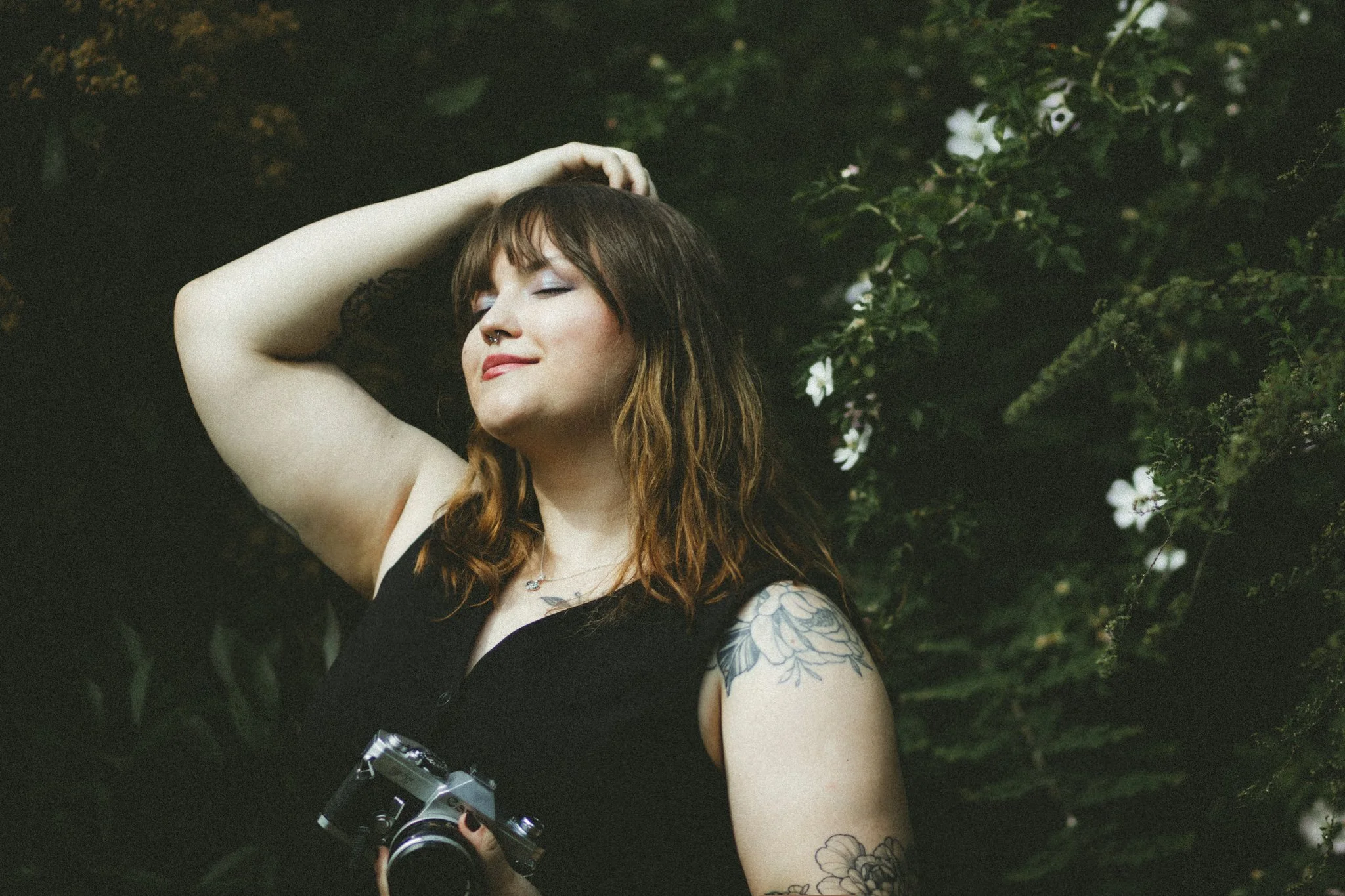 Young woman with tattoos, holding a vintage camera, smiling with eyes closed, standing outdoors near green foliage and white flowers.
