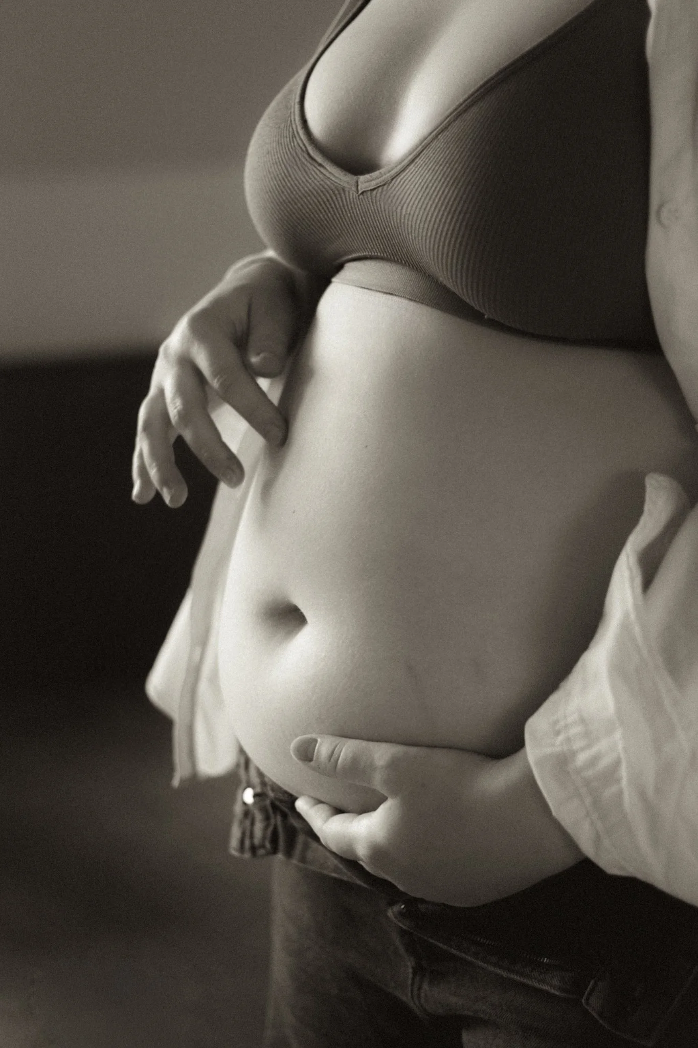 Close-up of a woman's torso and hands, wearing a sports bra and unzipped hoodie, with hands resting on her stomach in a black and white photograph.
