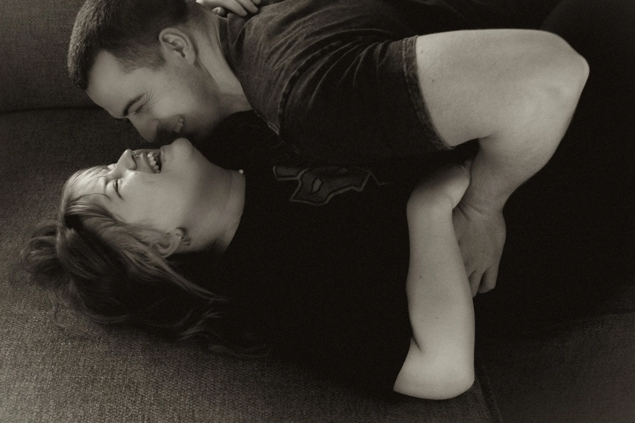 A young man and woman are lying on a couch, laughing and looking at each other closely in a joyful moment.