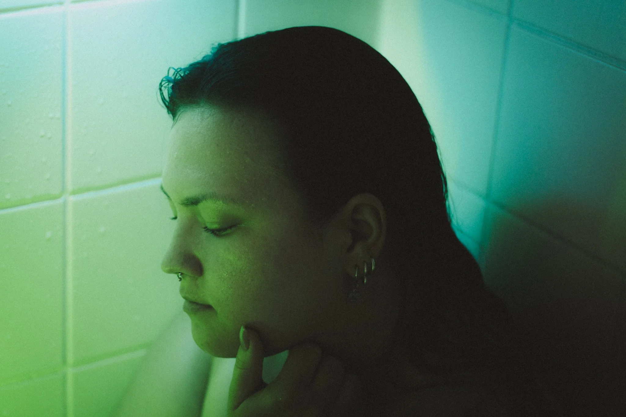 A person with dark, wet hair and earrings, standing against tiled wall with eyes closed, touching chin, illuminated by greenish light.