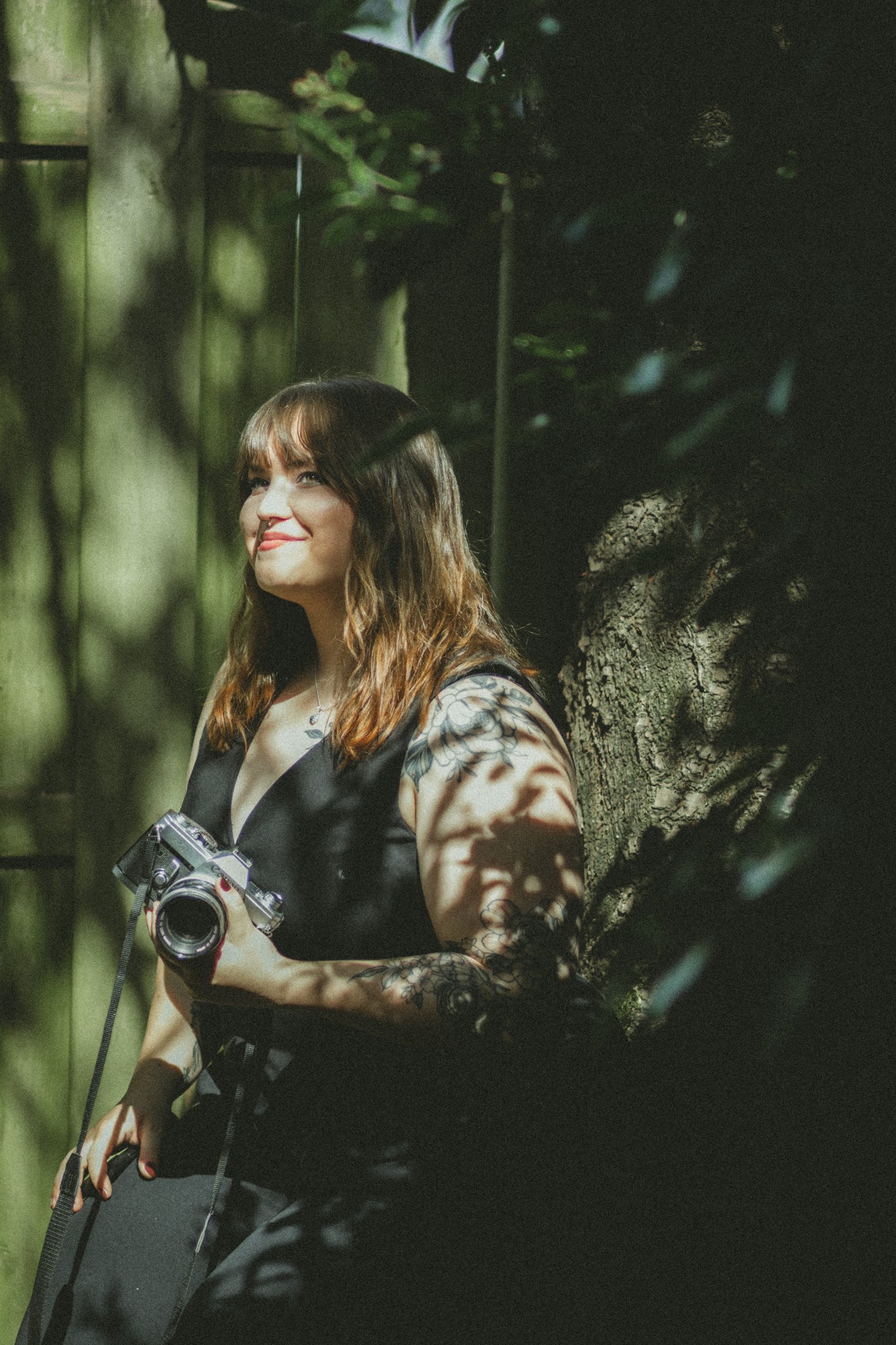 A woman with wavy brown hair and a tattoo on her arm holding a camera, standing beside a large tree trunk outdoors with dappled sunlight.