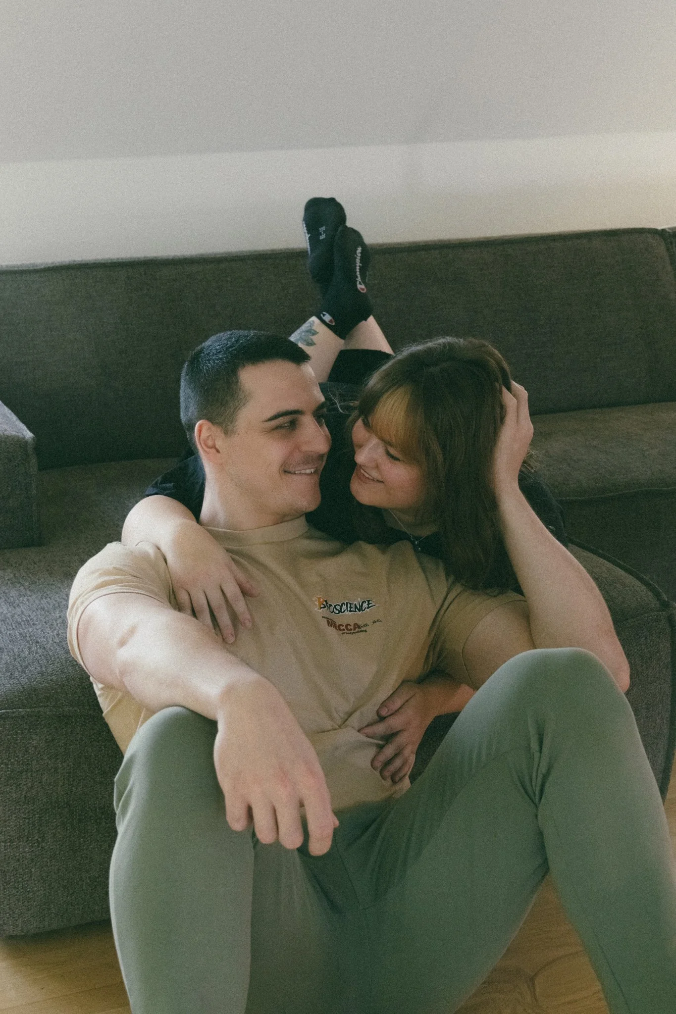 A couple sitting on the floor in front of a couch, smiling and embracing, with the woman leaning on the man's shoulder and his arm around her waist.