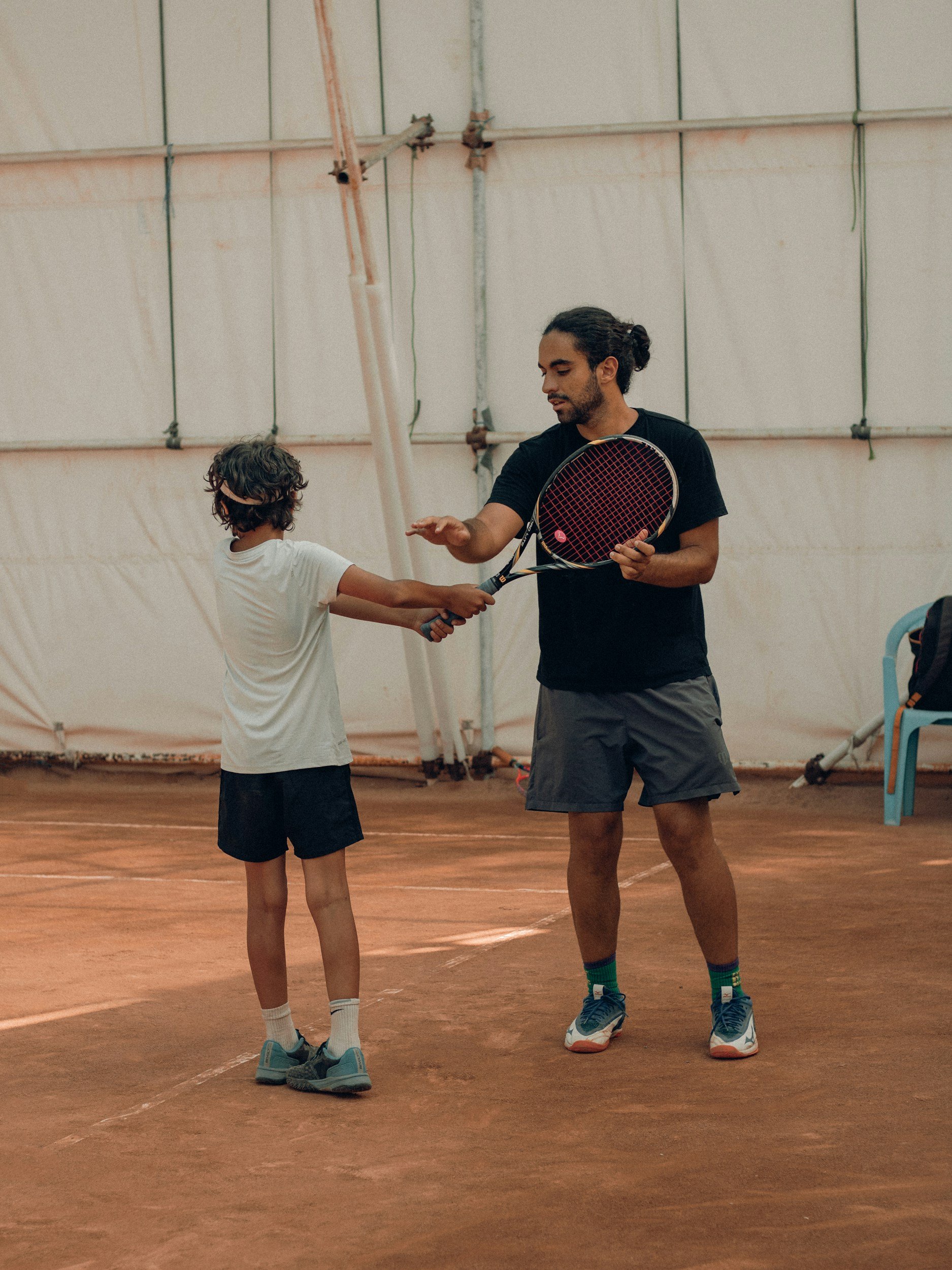 Tennis instructor teaching a young player on a clay court indoors.