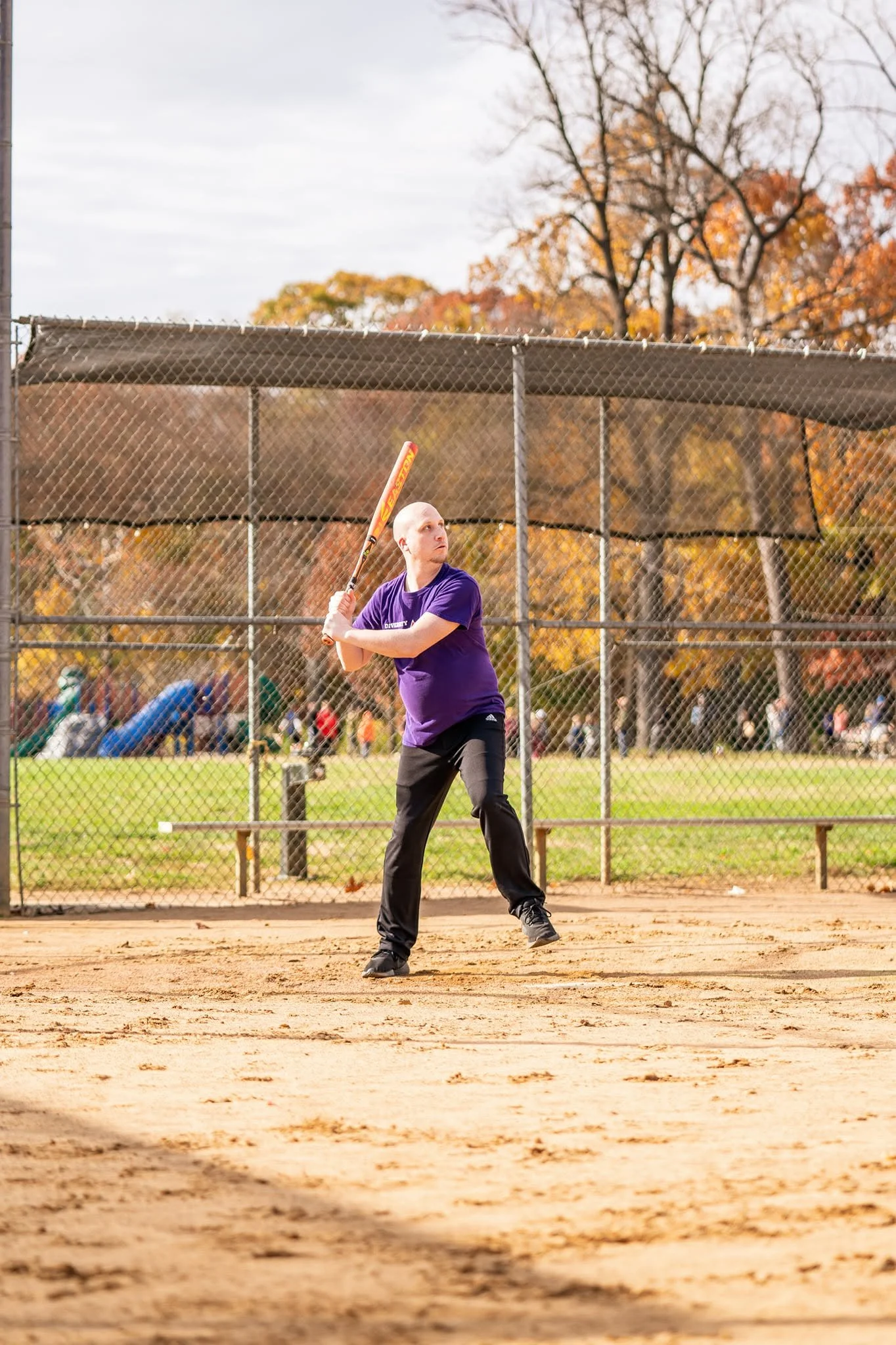 A person in a purple shirt and black pants standing on a baseball or softball field holding a bat, preparing to swing, with trees and a chain-link fence in the background during fall.