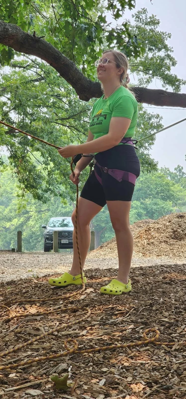 A young woman with glasses, wearing a green camp T-shirt, black shorts, bright yellow Crocs, and a harness, stands outdoors holding a rope near a large tree with green leaves. She appears to be preparing for a climbing or zip-lining activity, with a car parked in the background.