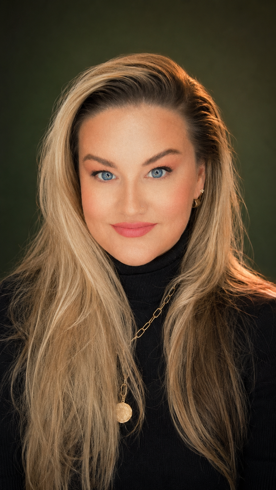A woman with long blonde hair, blue eyes, wearing a black turtleneck, gold necklace, and earrings, smiling against a dark green background.