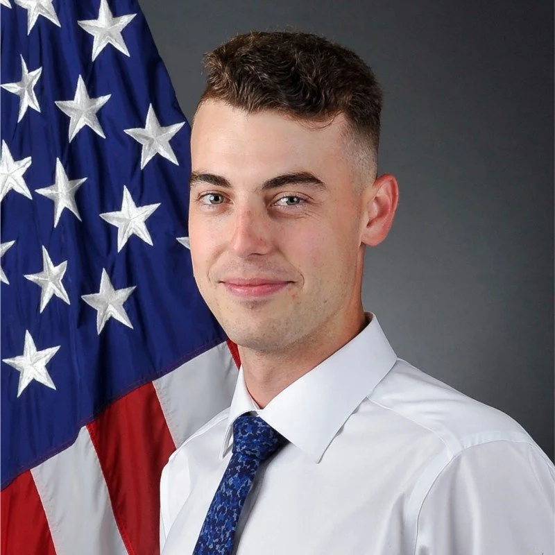 A young man in a white shirt and blue tie standing in front of an American flag.