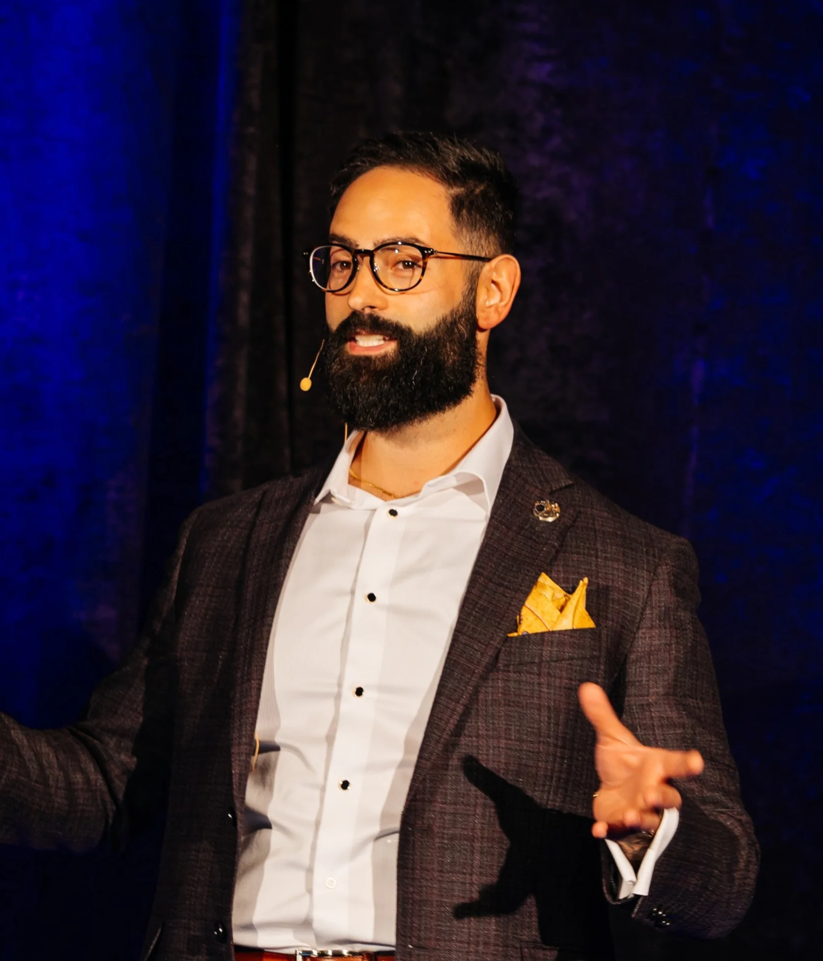 A man with dark hair, glasses, and a beard giving a presentation on stage. He is dressed in a checkered blazer, white shirt, and has a golden pocket square. He is wearing a headset microphone and gesturing with his hands in front of a dark backdrop with blue lighting.