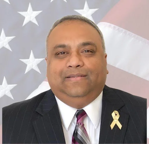 A man in a suit and tie with a gold ribbon pin, standing in front of an American flag background.