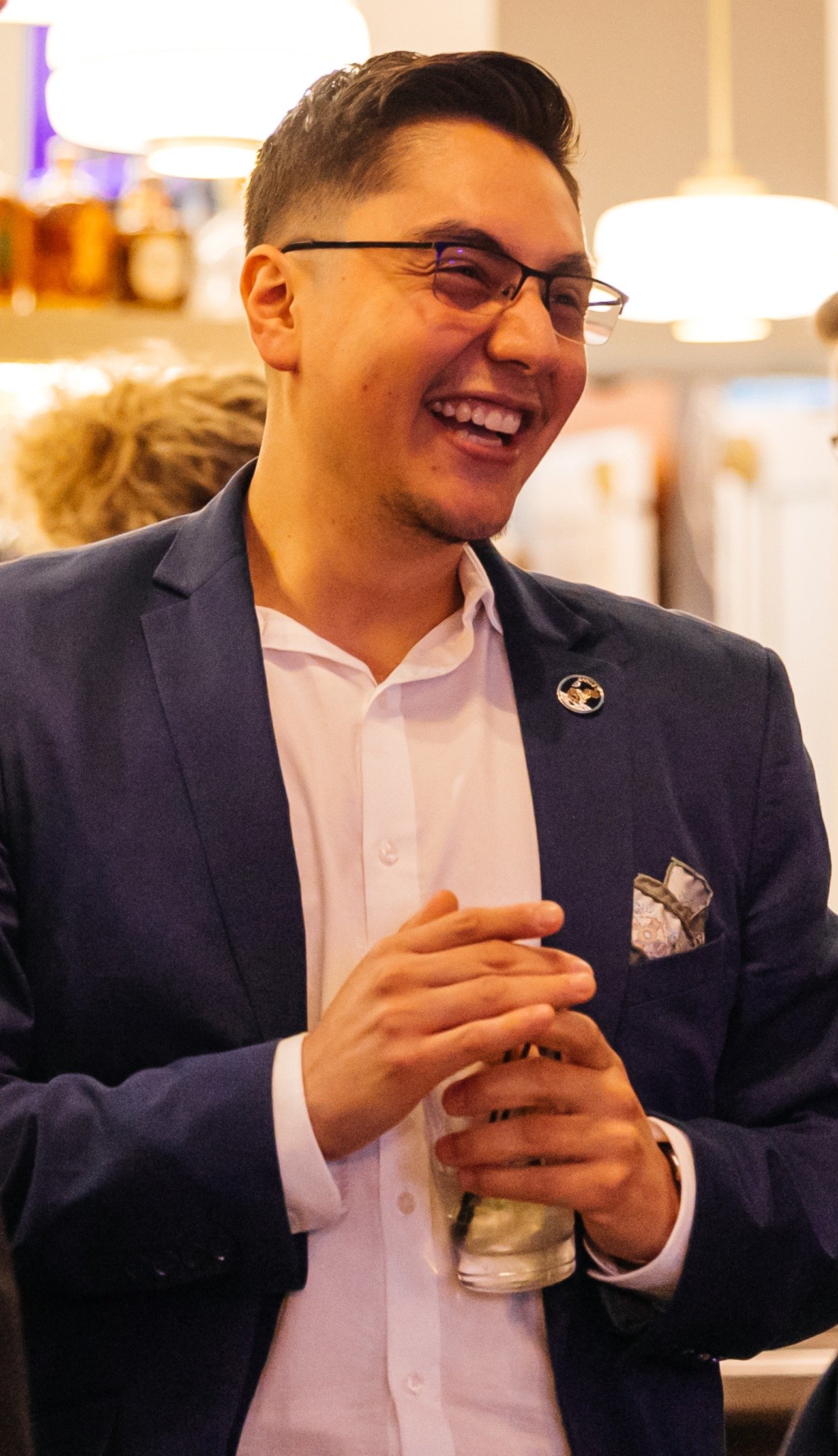 A young man wearing glasses in a dark suit, white shirt, smiling, holding a drink, at a social gathering in a warmly lit room.