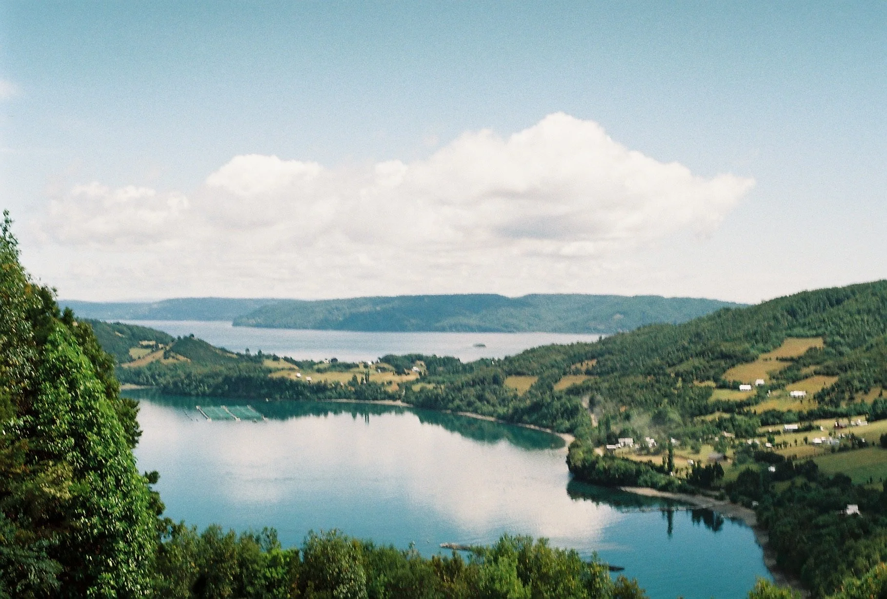 Scenic view of a river flowing through green hills with scattered houses and trees, under a partly cloudy sky.