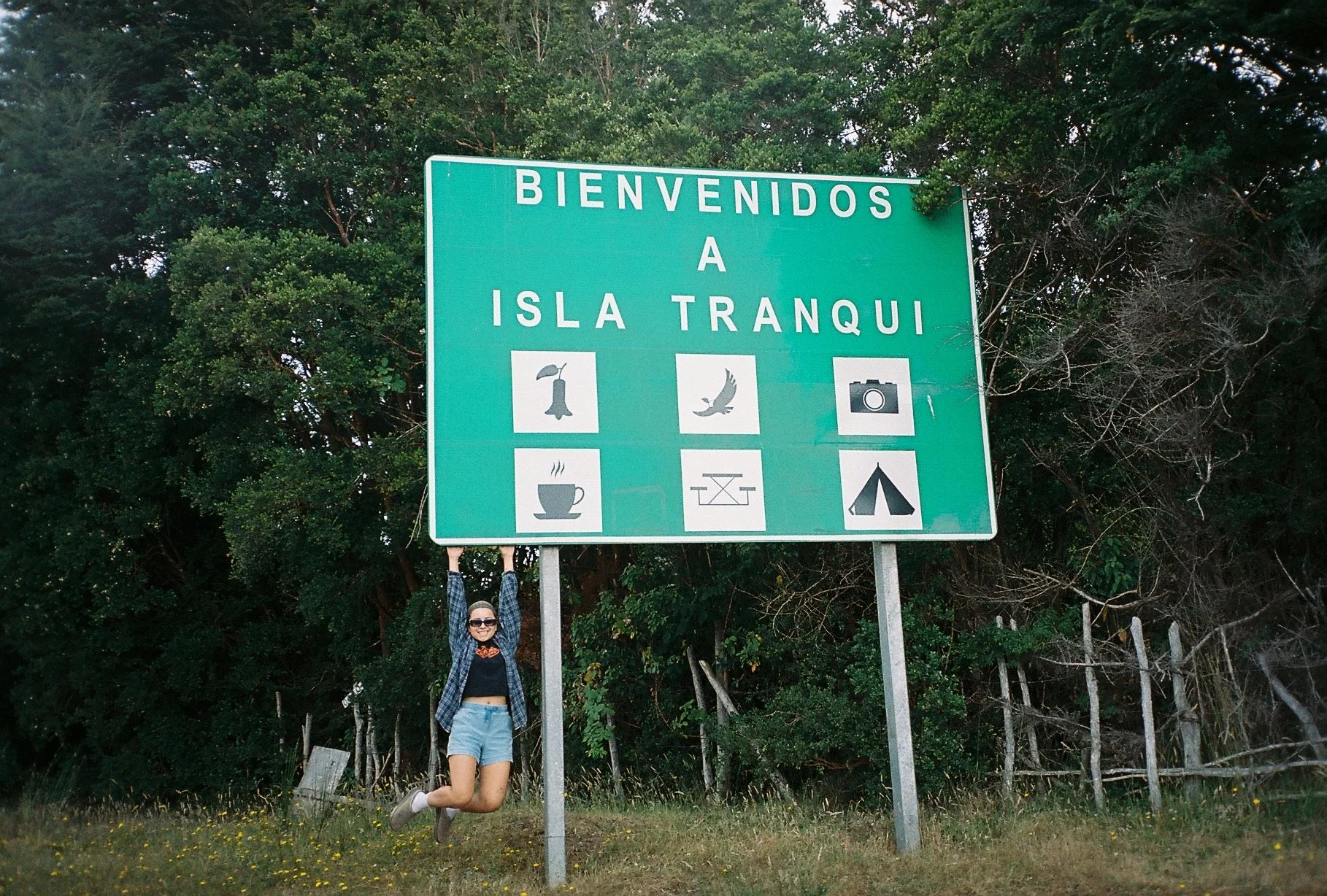 Woman with sunglasses smiling, wearing a plaid shirt and shorts, hanging from a road sign that says 'Bienvenidos a Isla Tranquila' against a background of trees and a partly cloudy sky.
