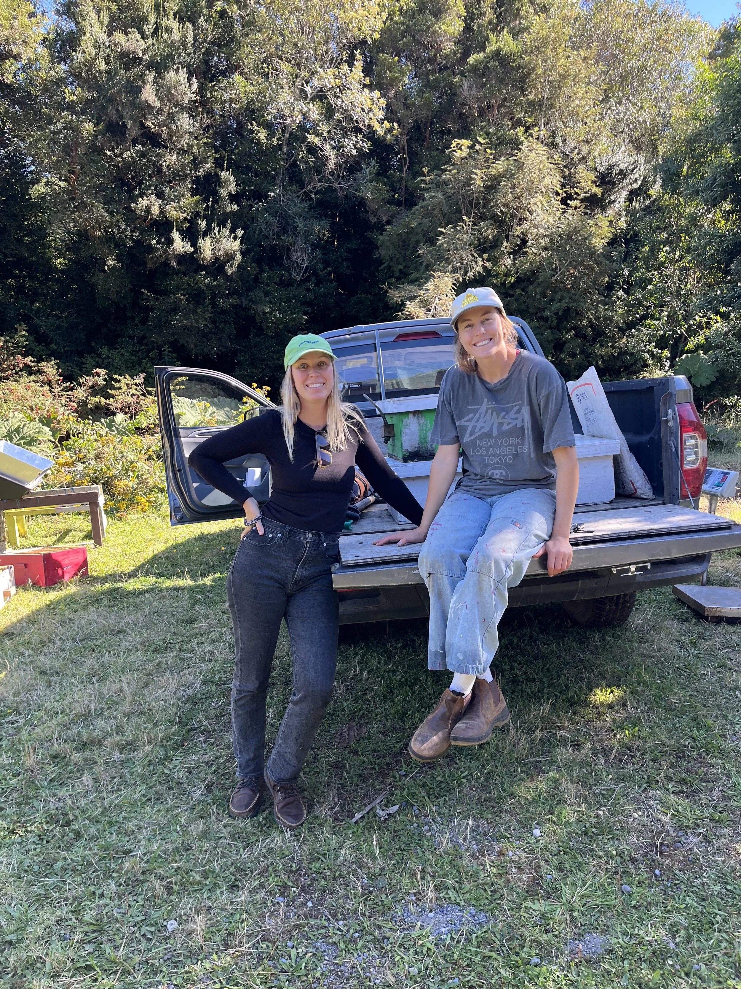 Two women sitting and standing in front of an open truck bed outdoors, with a dense green tree line in the background. They are smiling and appear to be working or gardening with gardening supplies nearby.