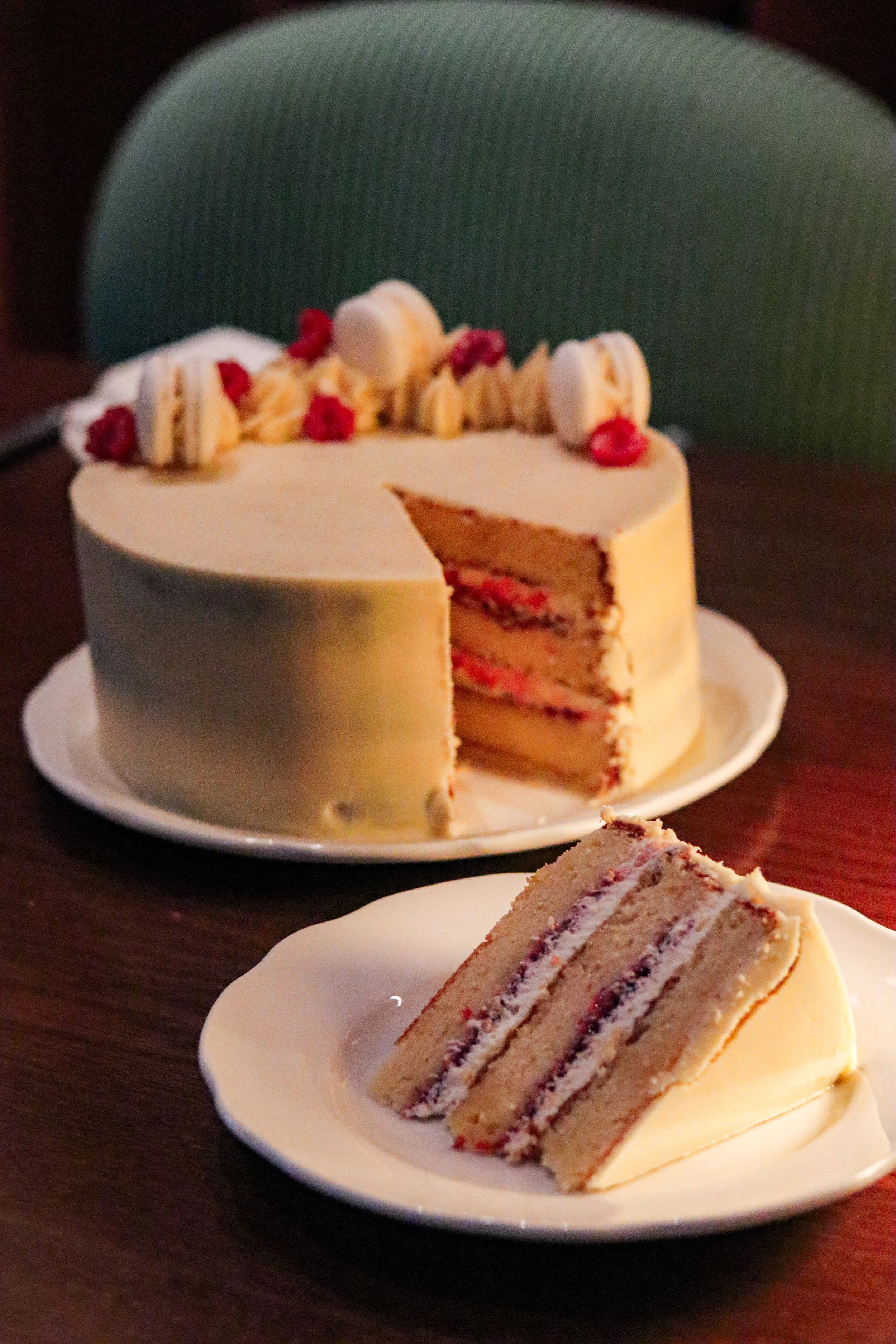 A round cake with white frosting on a white plate, with a slice removed showing layers of white cake and pink filling, topped with macarons, raspberries, and cream dollops. A slice of the cake is on a separate white plate in front.