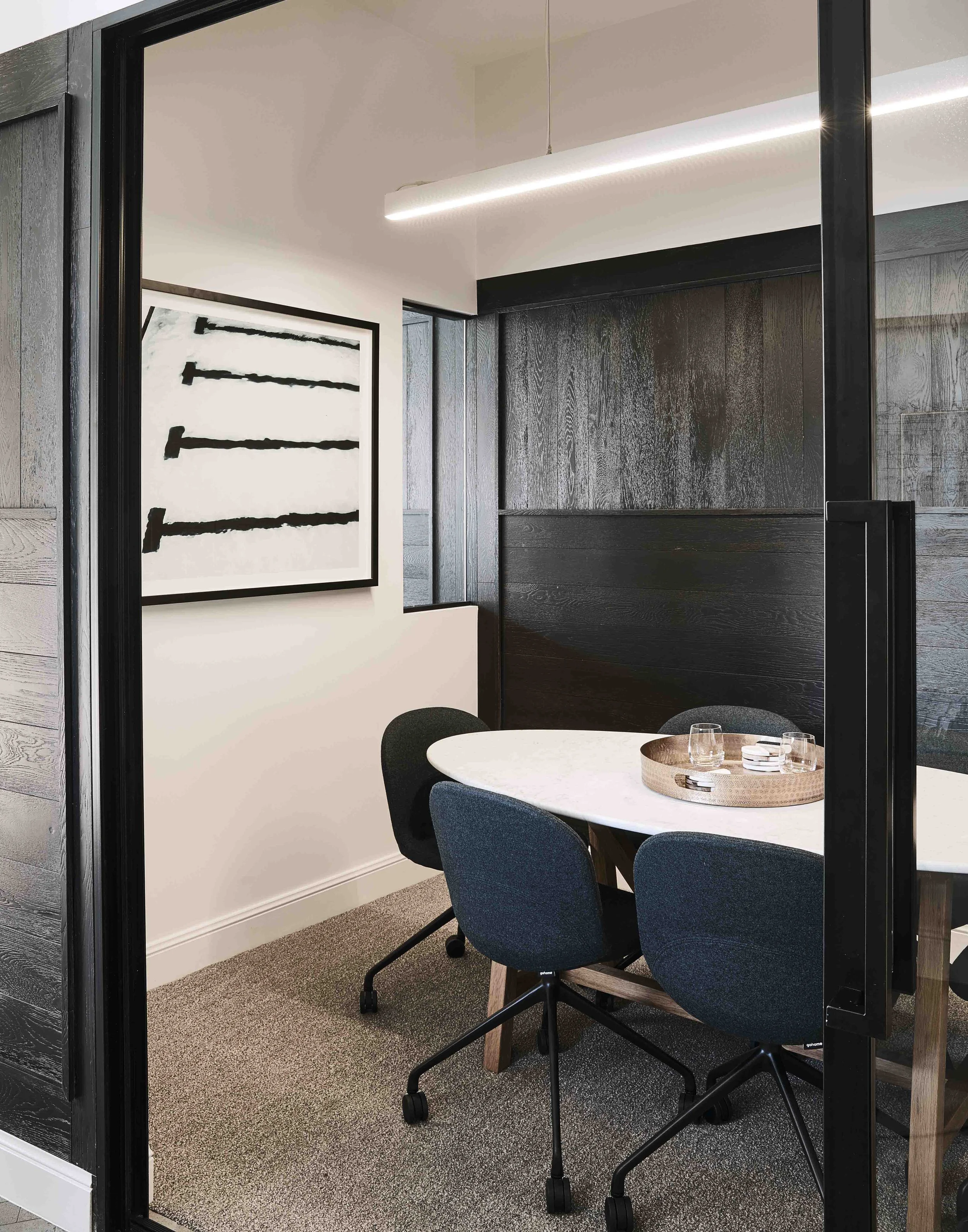 Modern conference room with white oval table, four black and blue office chairs, black wood-paneled wall, abstract black and white artwork, and a long ceiling light.