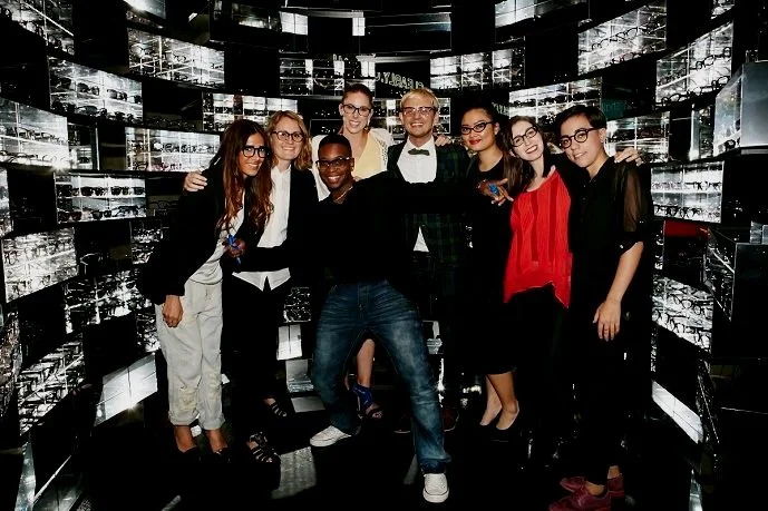 A group of eight young people, mostly women and one man, smiling and posing together inside a high-end watch display room with reflective walls.