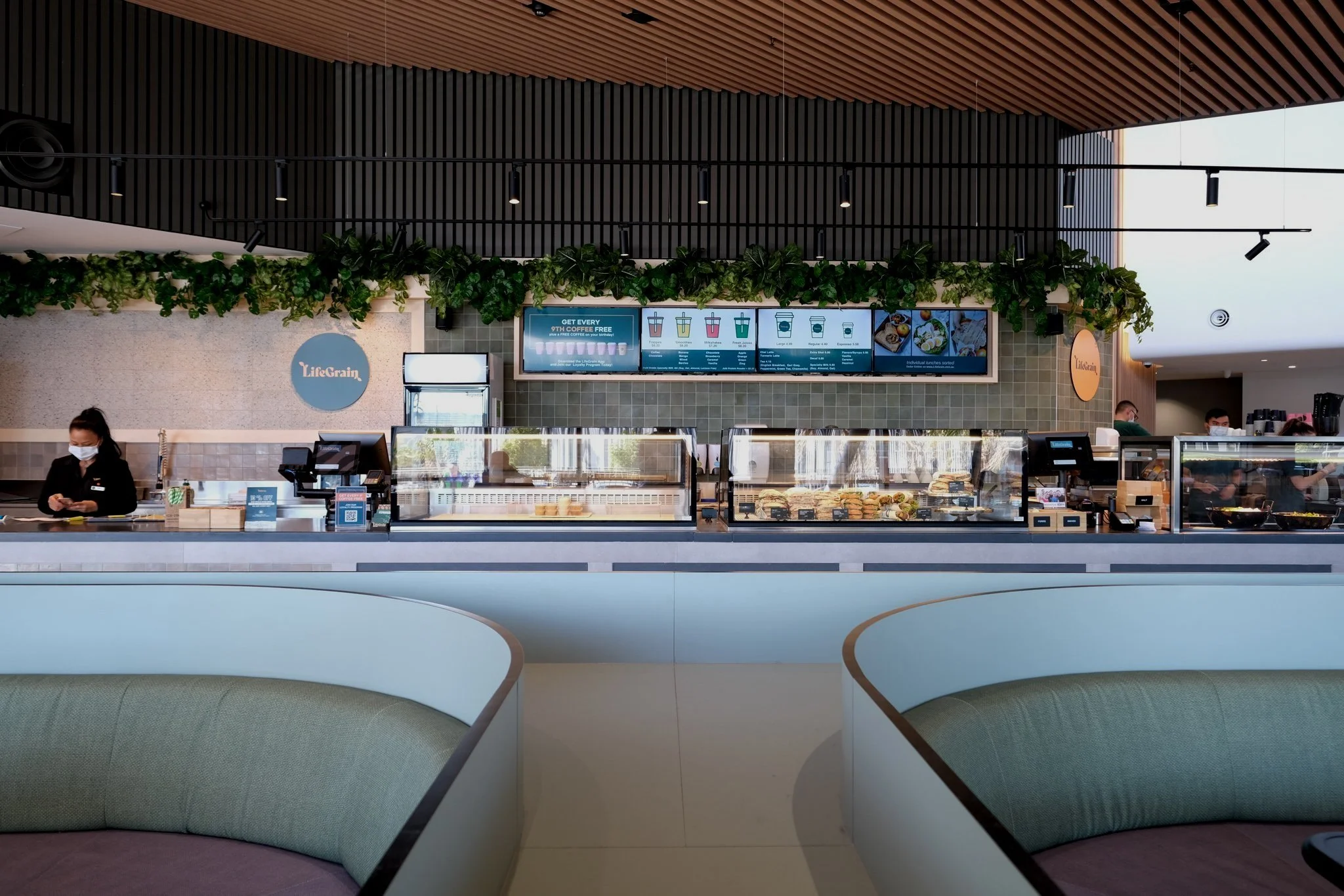 Inside a modern coffee shop with a counter displaying baked goods, a Barista wearing a mask working, digital menu screens above, and seating area in the foreground.