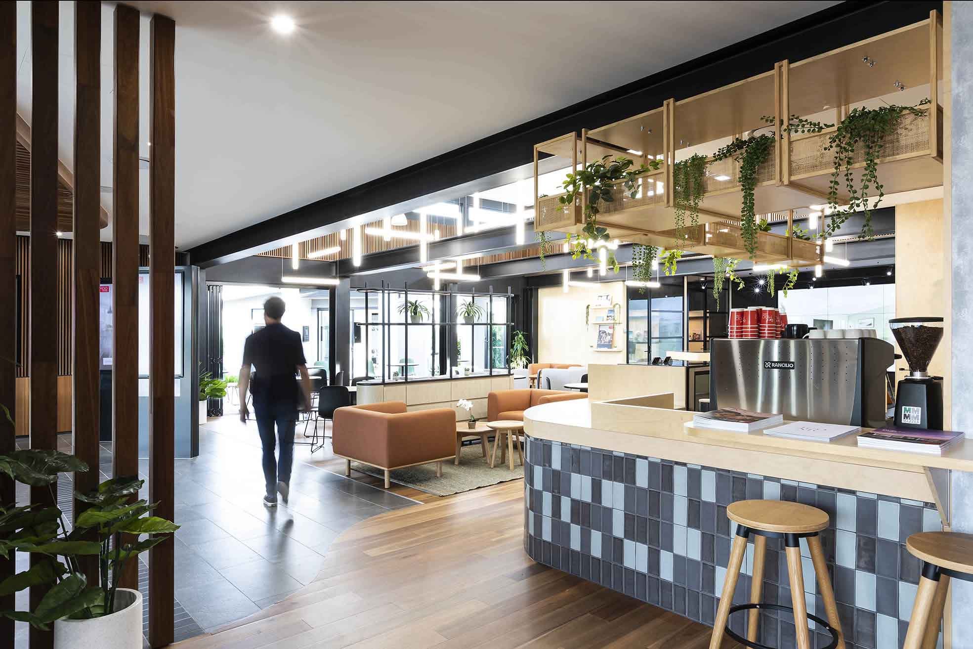 Modern café interior with seating areas, café counter with a coffee machine, and decorative greenery, illuminated by natural and artificial light.