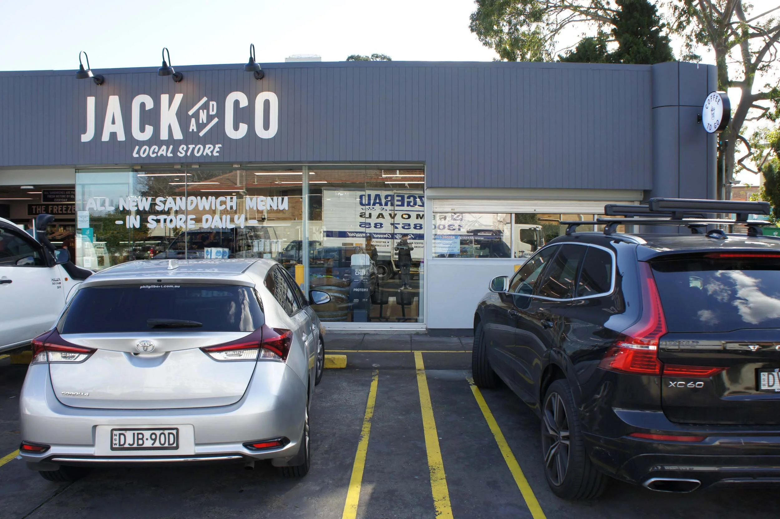 Front view of Jack and Co local store with parked cars in the parking lot, including a silver Toyota and a black Volvo XC60, and a sign advertising a new sandwich menu.