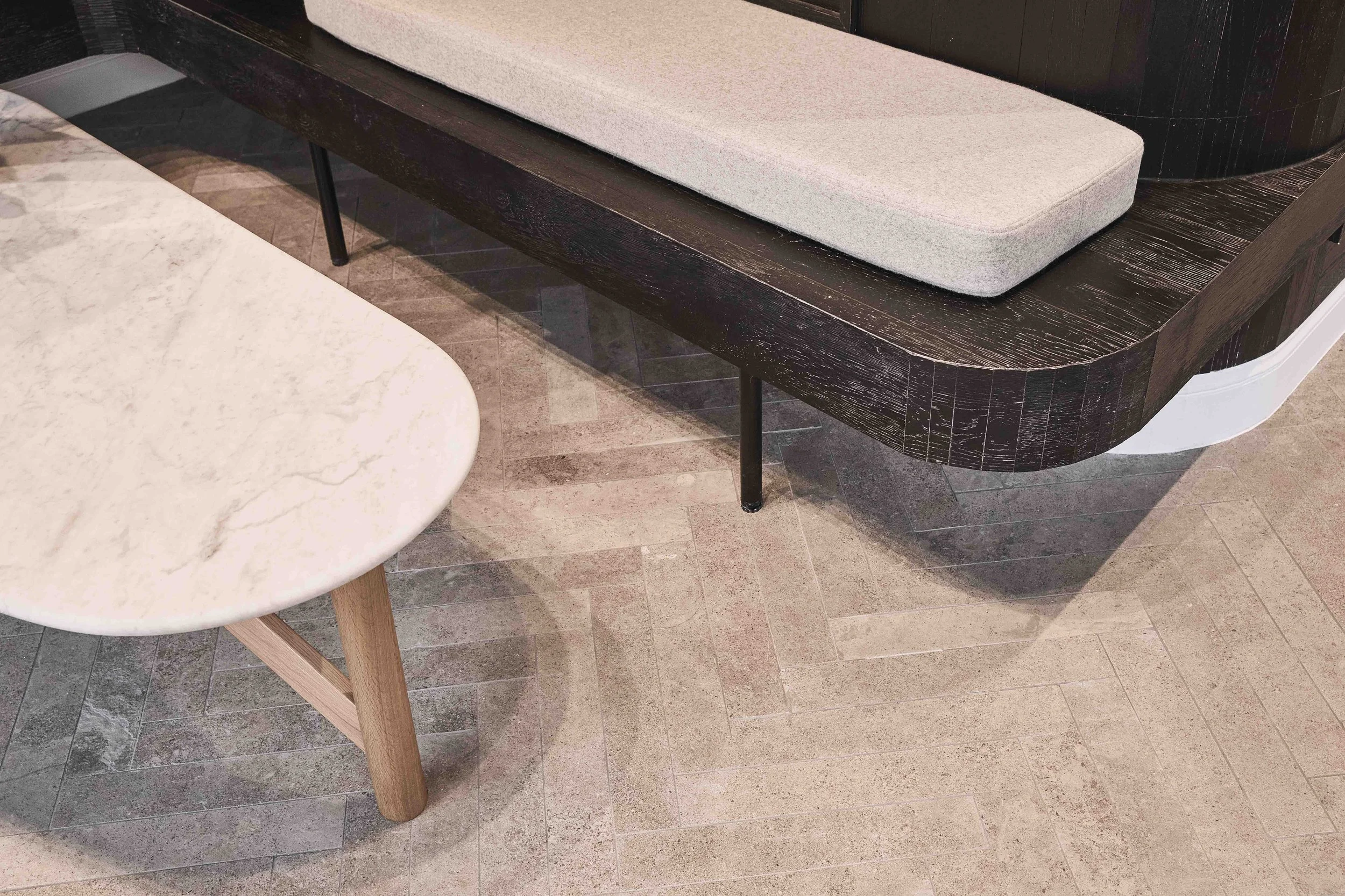 A close-up view of a marble table with wooden legs, a dark wood bench with a beige cushion, and a tiled floor in a modern interior setting.