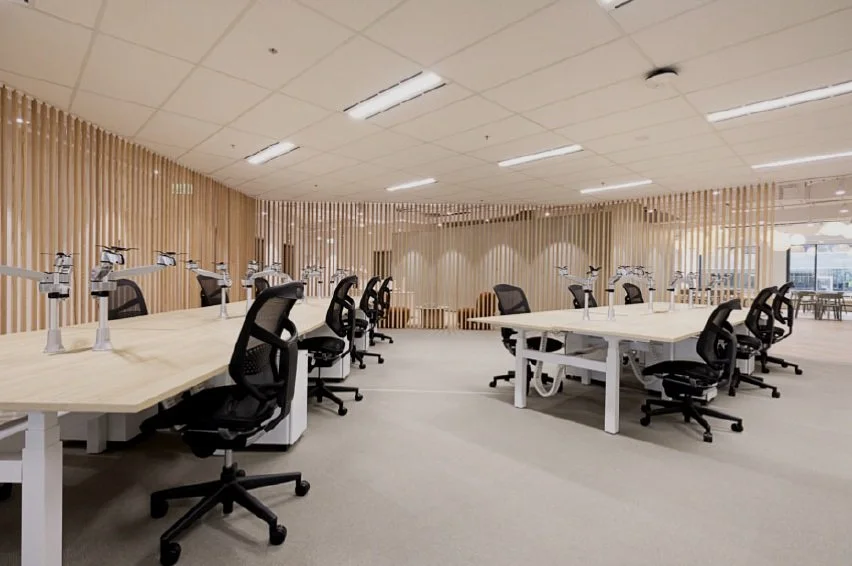 Modern office workspace with multiple ergonomic chairs and white desks, illuminated by ceiling lights, featuring wooden slat partition walls.