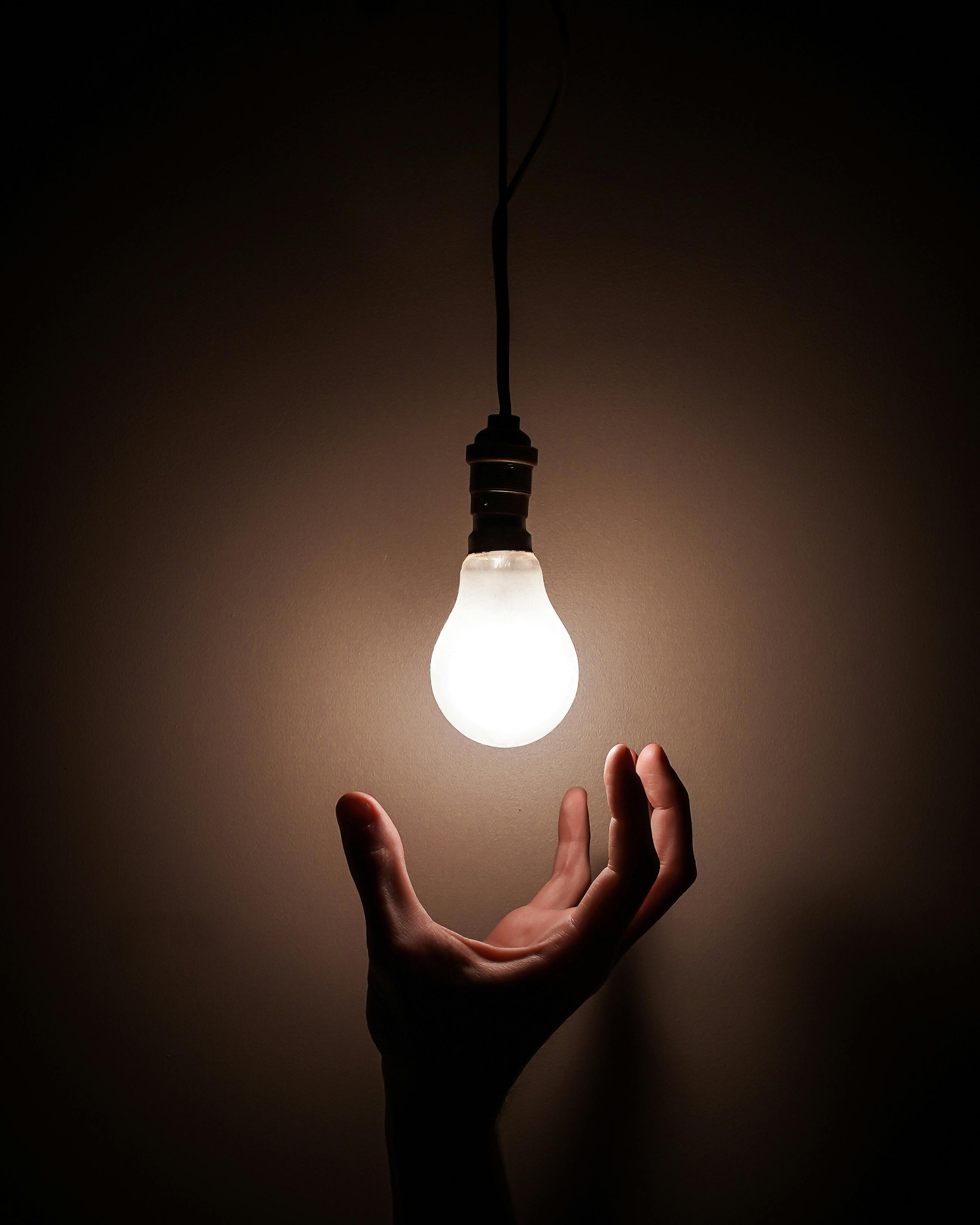 A hand reaching towards a lit light bulb hanging from the ceiling.