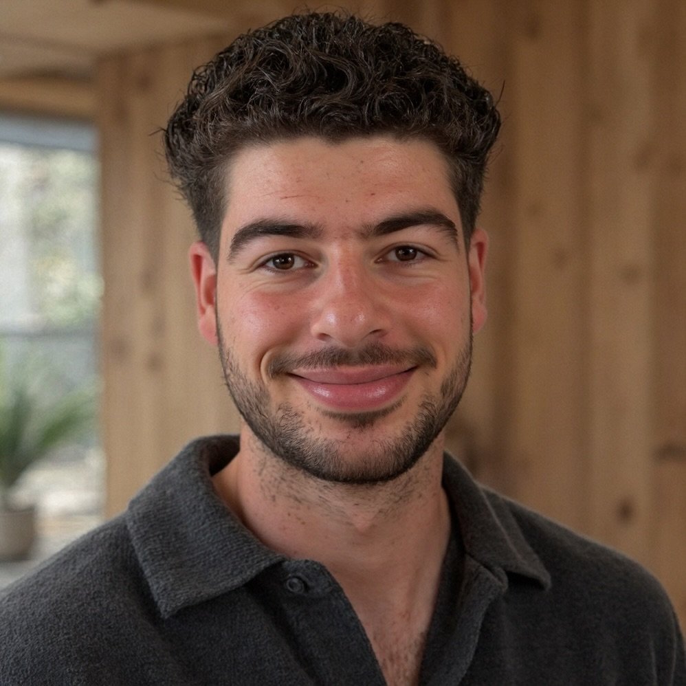 Smiling man with curly hair and a beard standing indoors with wooden background.