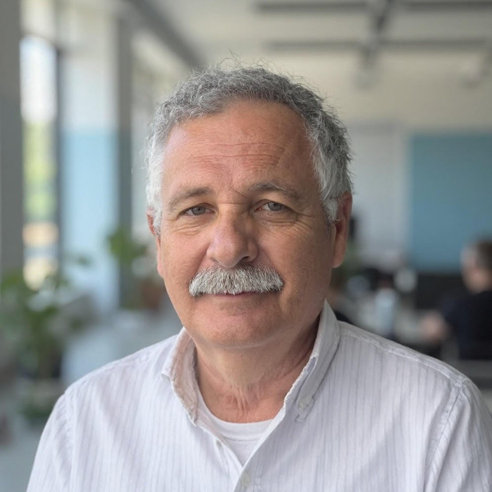 Elderly man with gray hair and mustache wearing a white shirt in a light-filled office space.