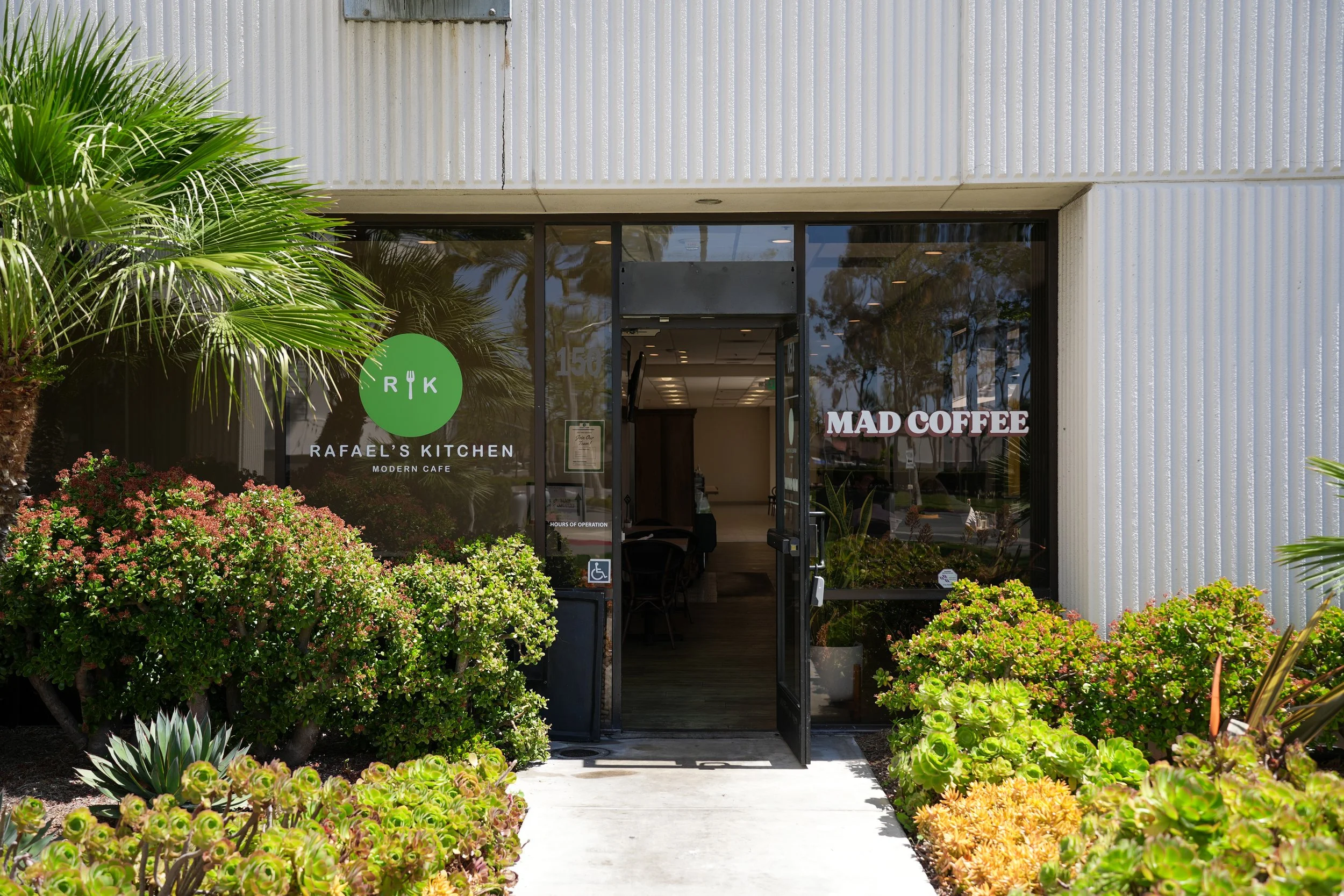 Entrance of Rafael's Kitchen, a modern cafe with glass doors, surrounded by lush green plants and bushes, with signs for Mad Coffee visible on the window.