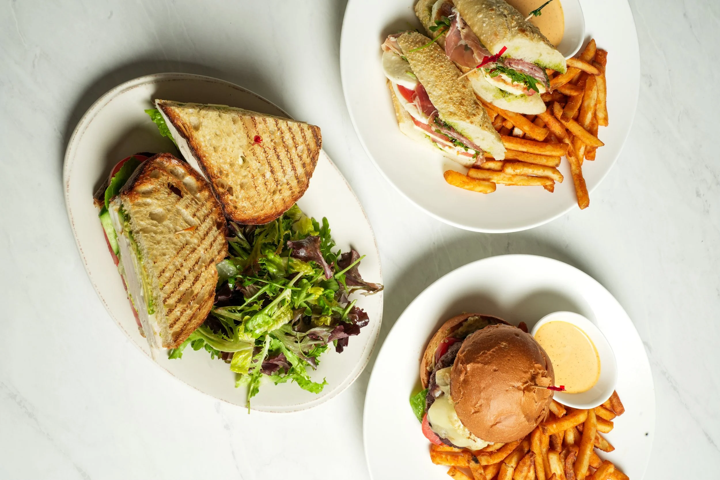 Top view of three plates with sandwiches and fries, and a side salad on a white surface.